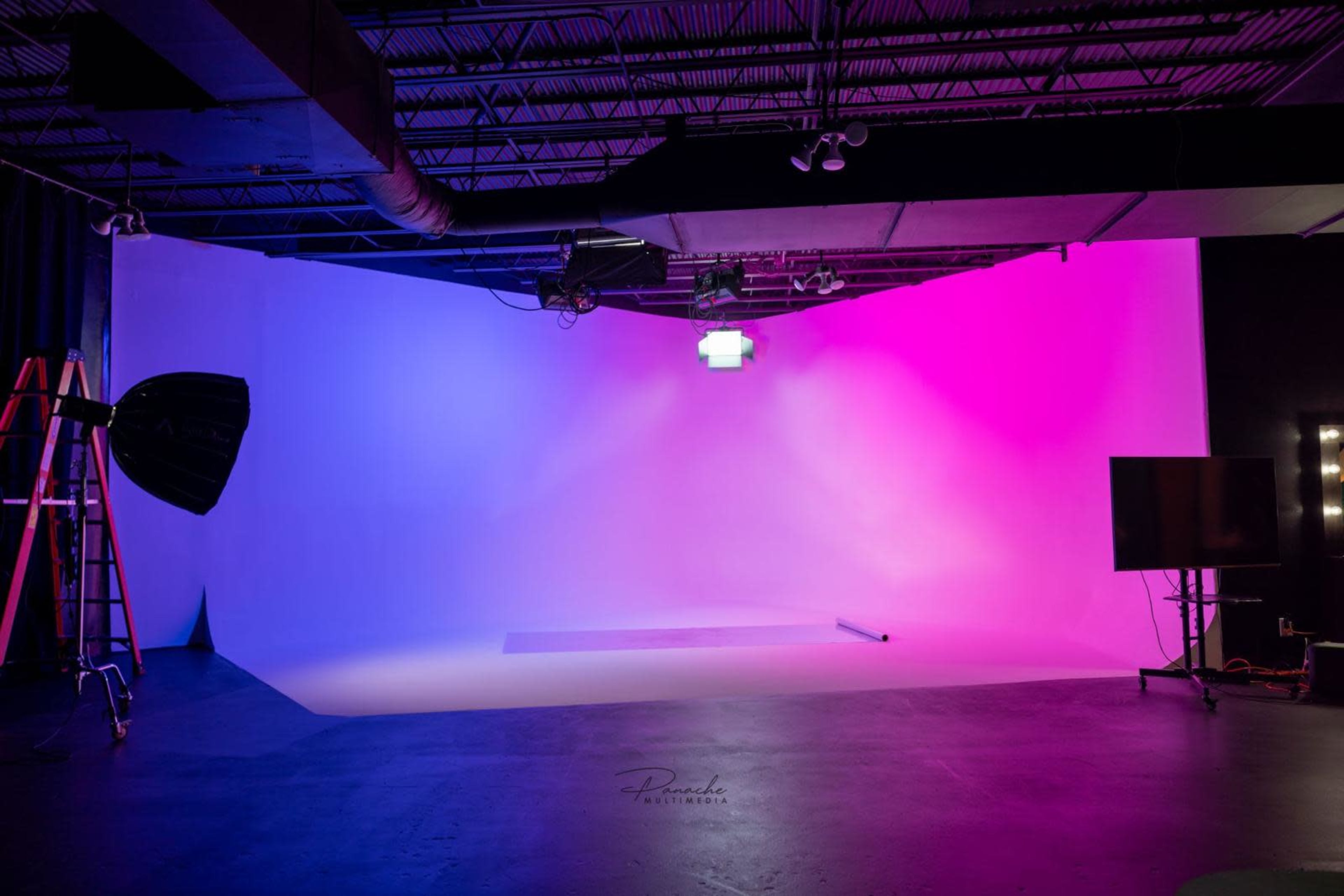A photography studio with a gradient backdrop featuring purple and blue lighting, a light stand, a ladder, and a television monitor.