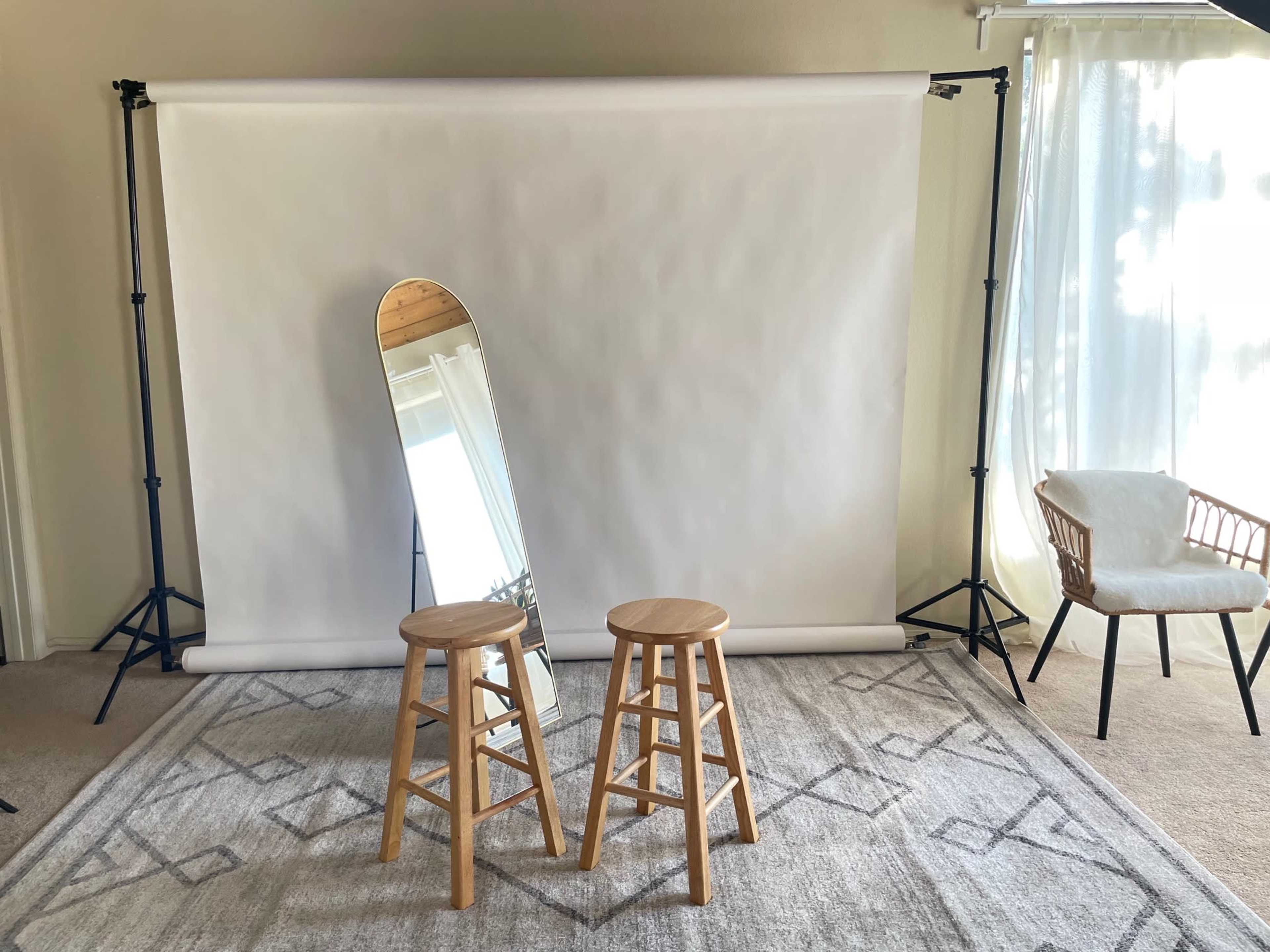The image shows a simple indoor setup with a blank white backdrop, two wooden stools, an arched mirror, and a chair beside a window covered with sheer curtains.
