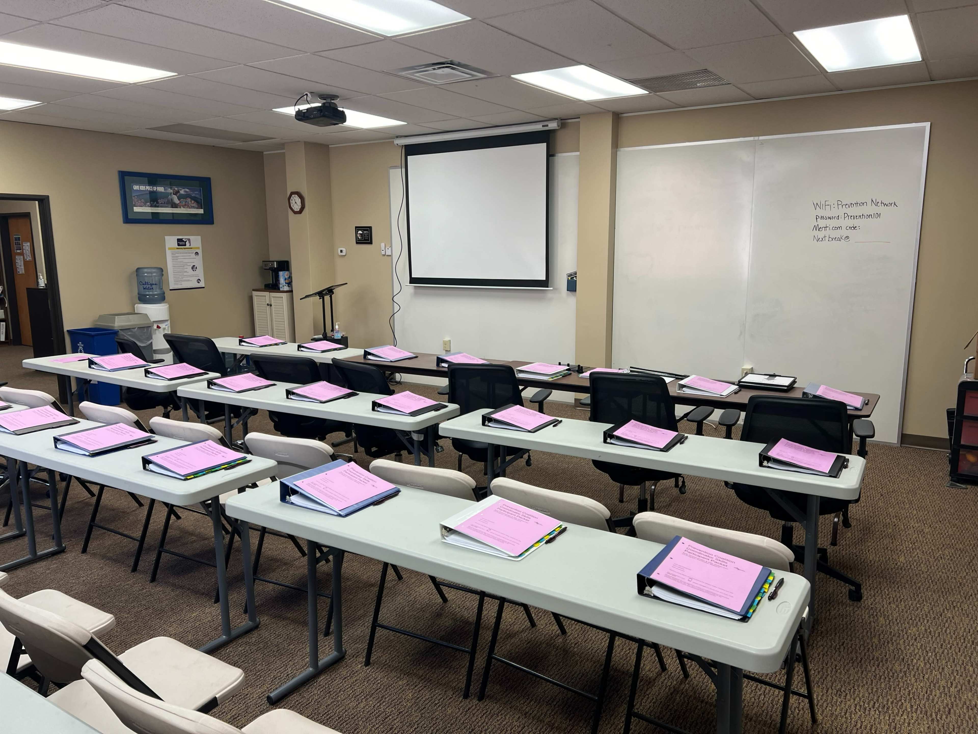 A classroom is set up with rows of tables and chairs, each holding a pink folder, and a projector screen is displayed at the front.