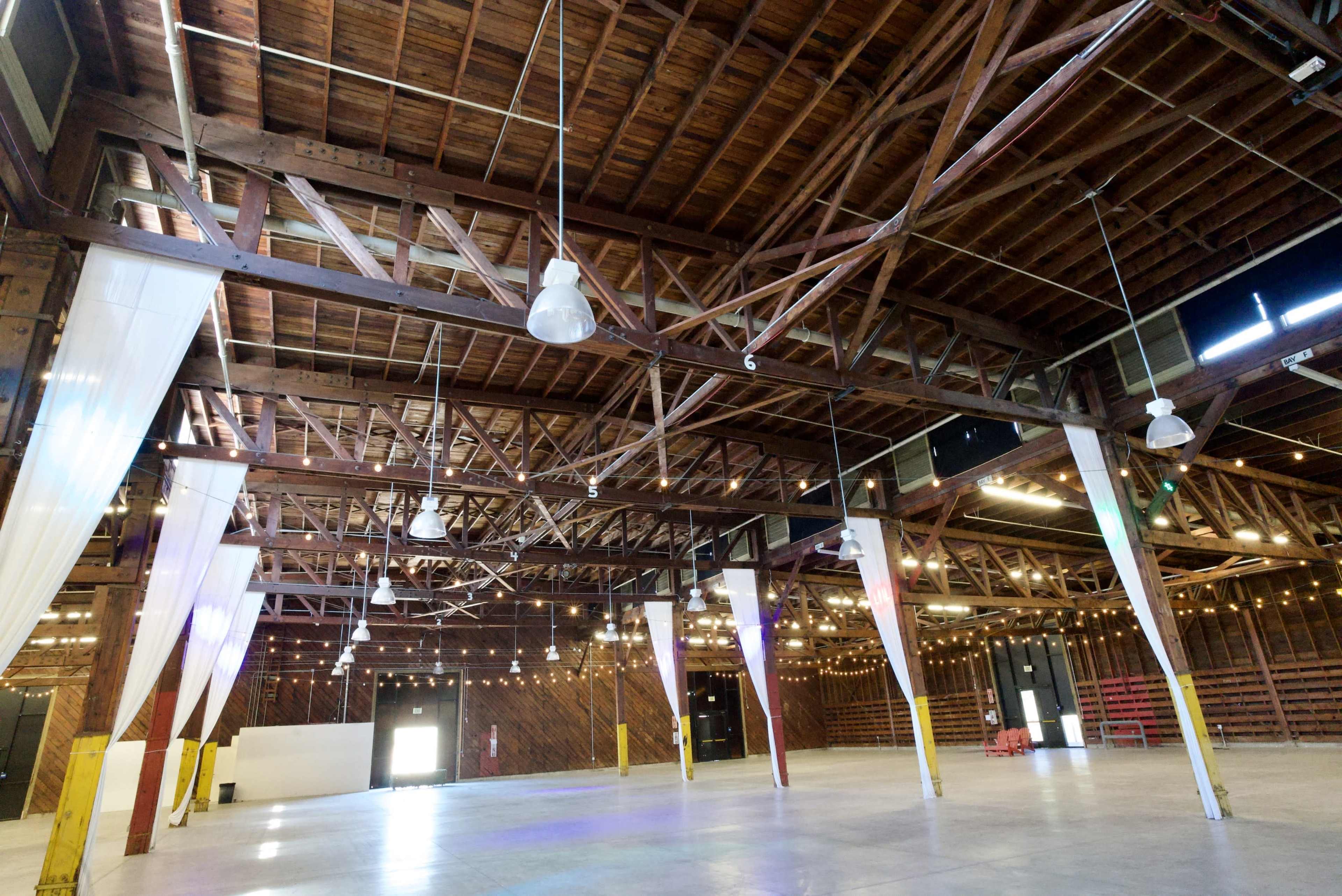 The image shows a spacious interior of a renovated barn with exposed wooden beams, industrial lights, and decorative string lights hanging from the ceiling.