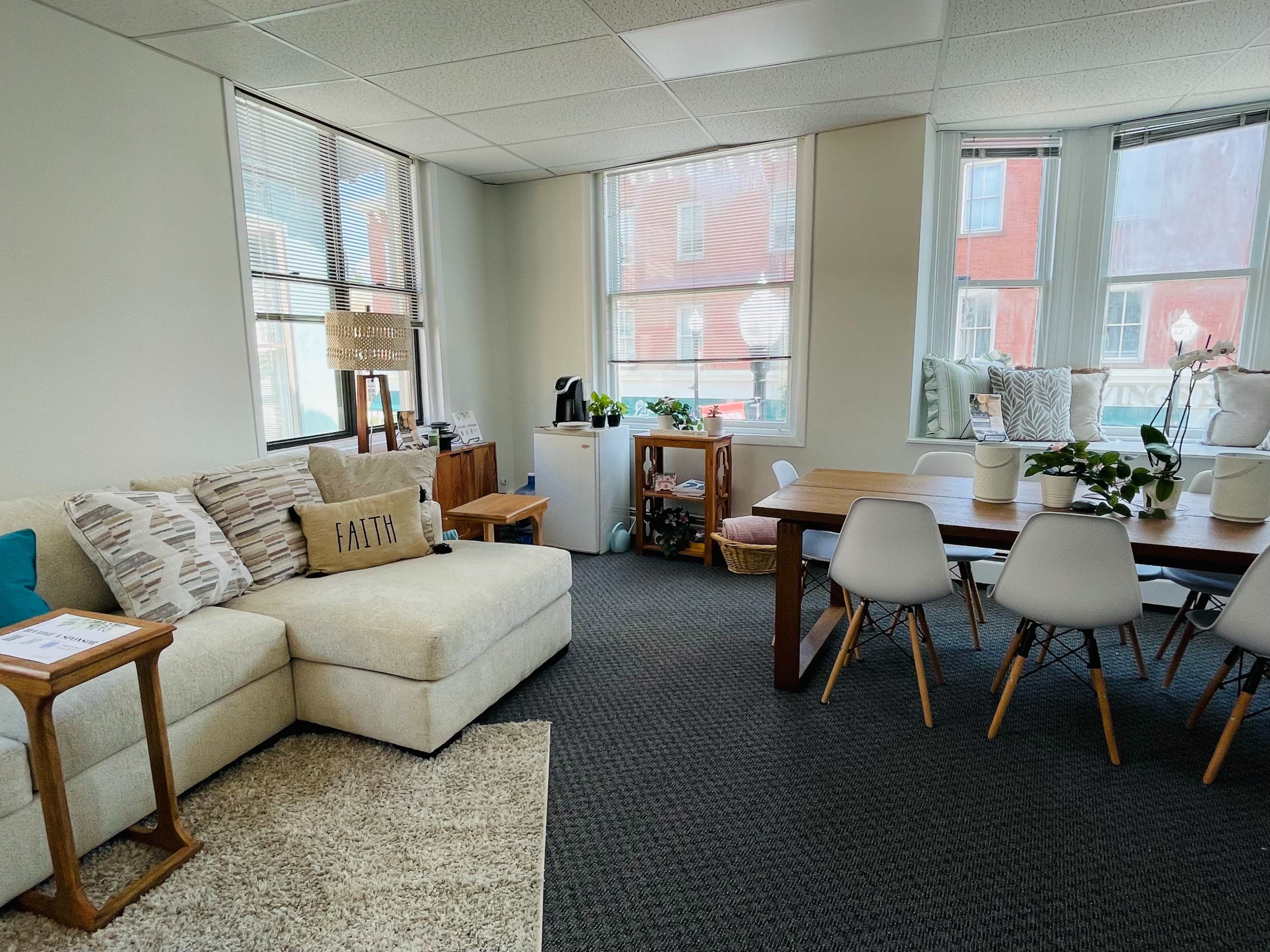 The room features a beige sectional sofa, a wooden dining table with white chairs, and large windows that provide natural light.