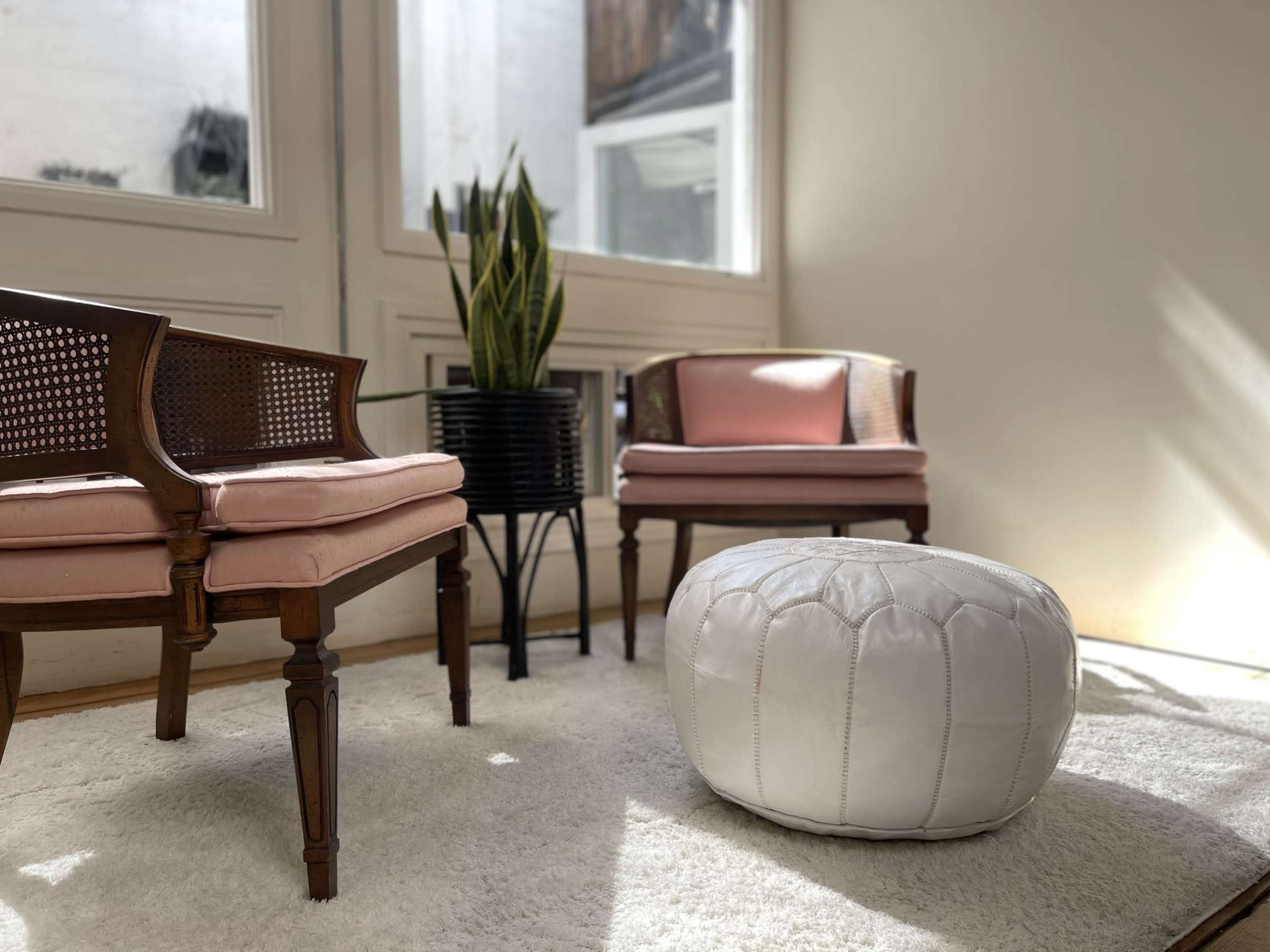 Two pink upholstered chairs and a white pouf arranged on a light rug in a well-lit room with a plant in a black pot.