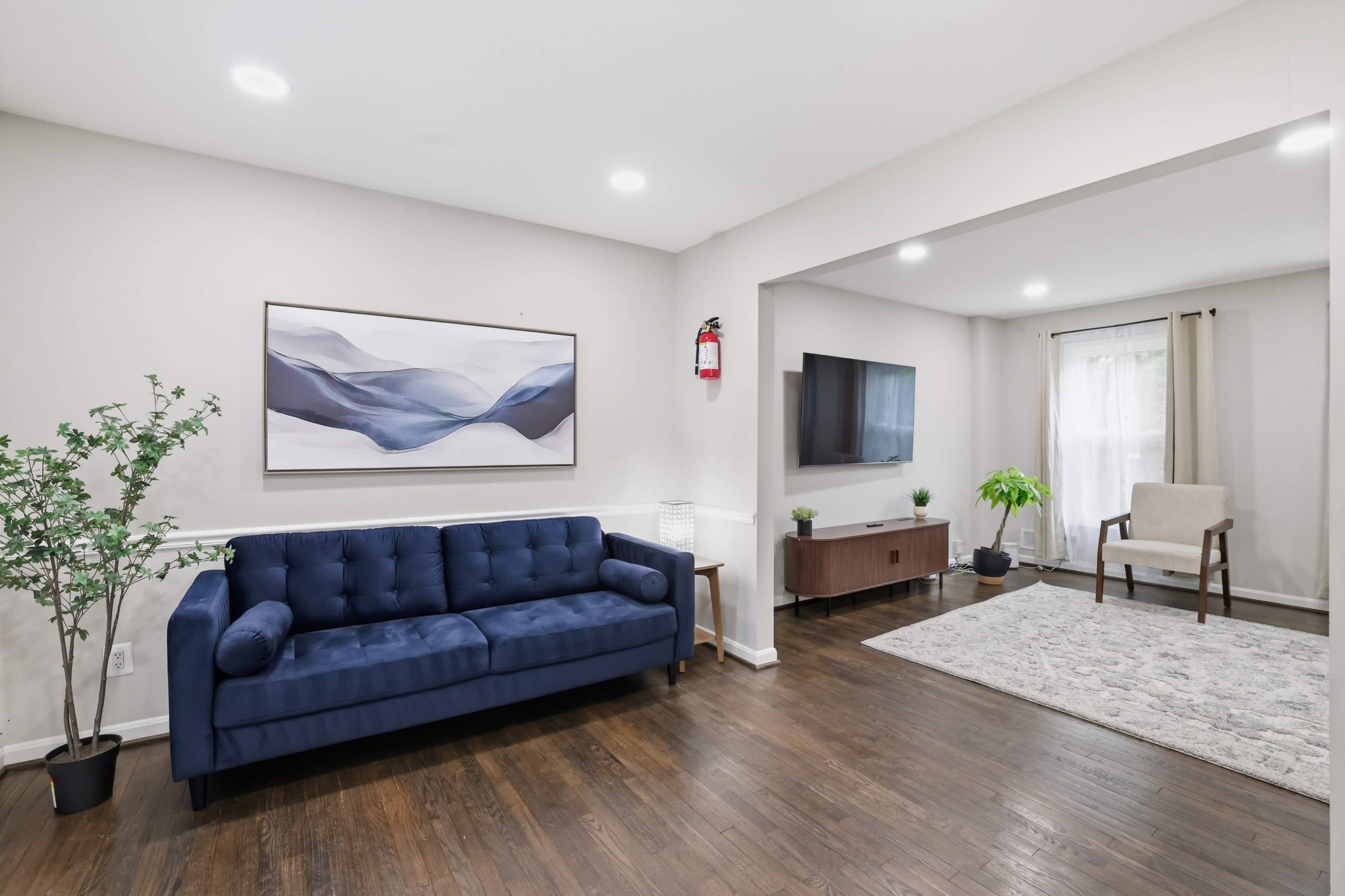 The image shows a modern living room with a blue sofa, a television mounted on the wall, a small wooden entertainment unit, and a rug on the wooden floor.