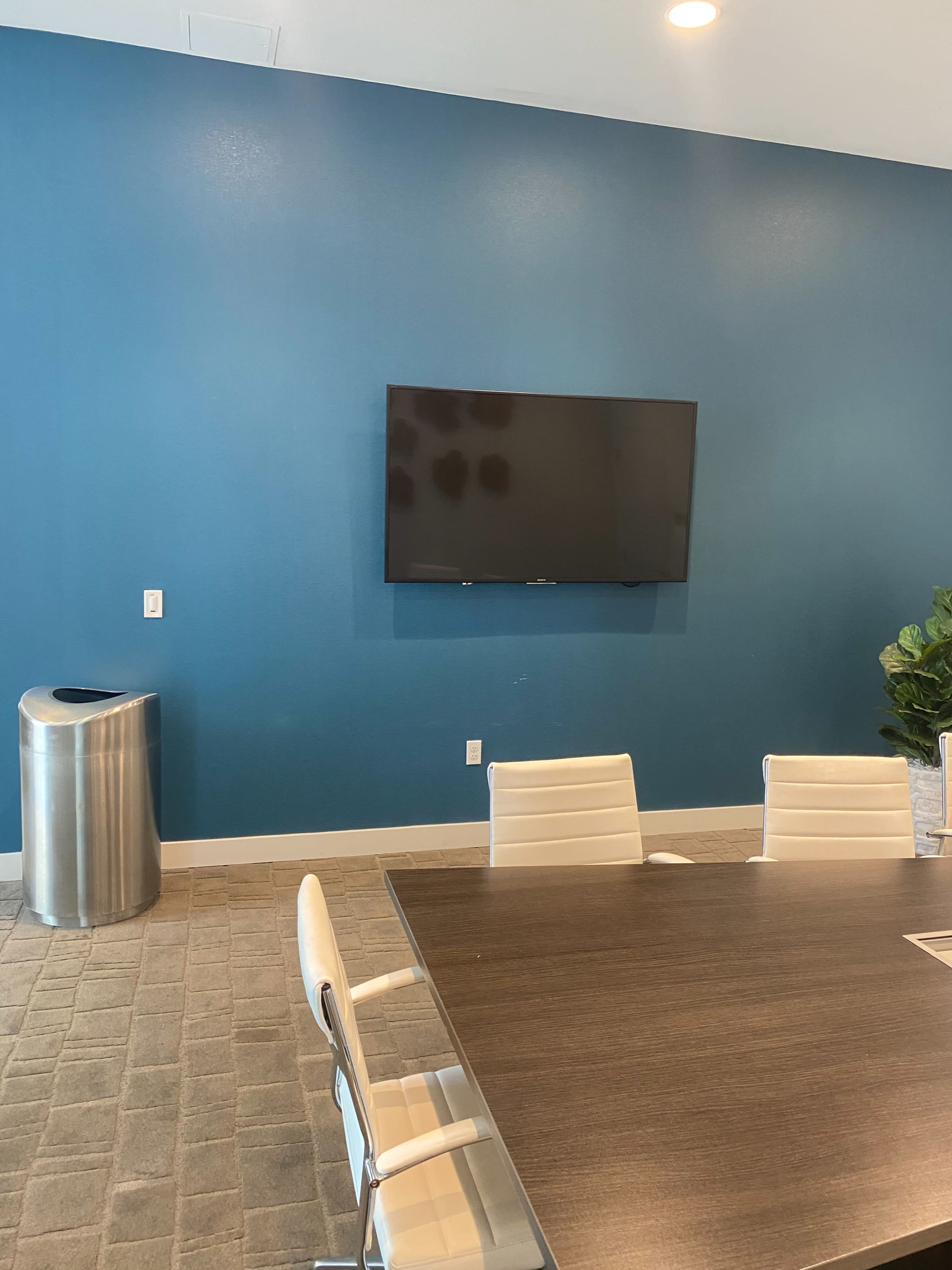 A conference room features a large table surrounded by white chairs, a mounted television on a blue wall, and a metal trash can in the corner.
