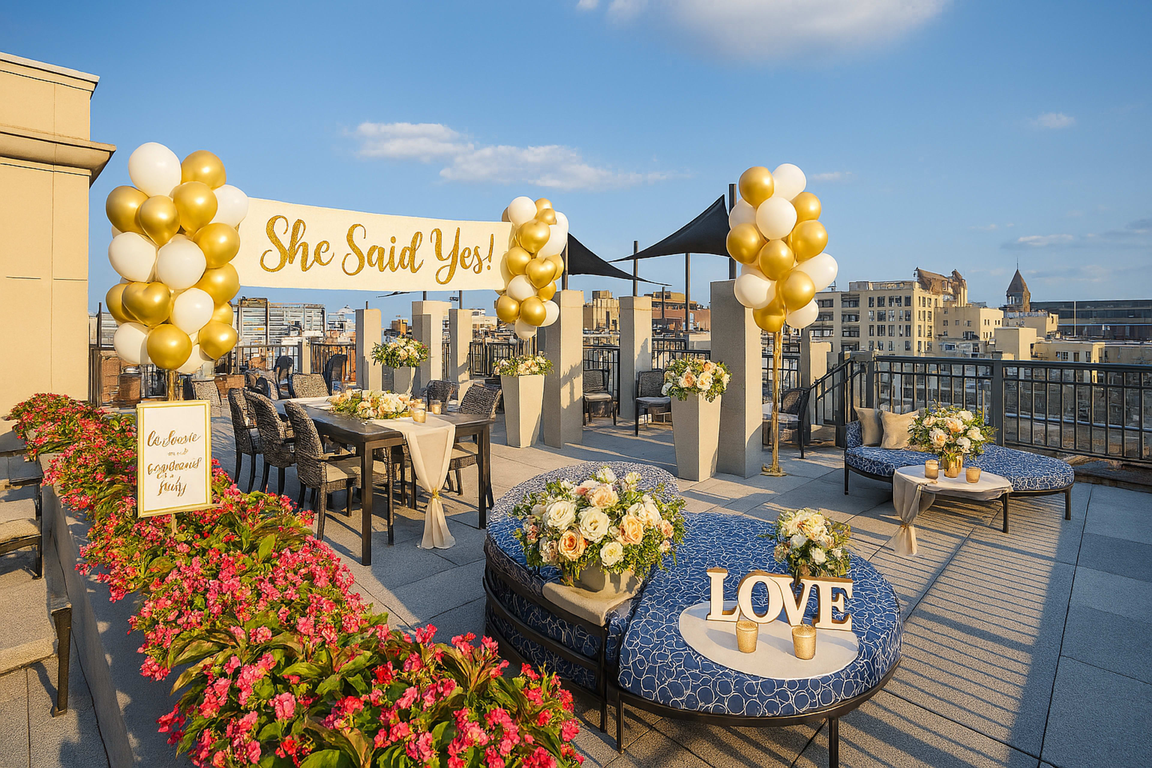 The image shows a rooftop celebration setup with decorated seating, floral arrangements, and a large banner that reads "She Said Yes!" amidst a clear blue sky.