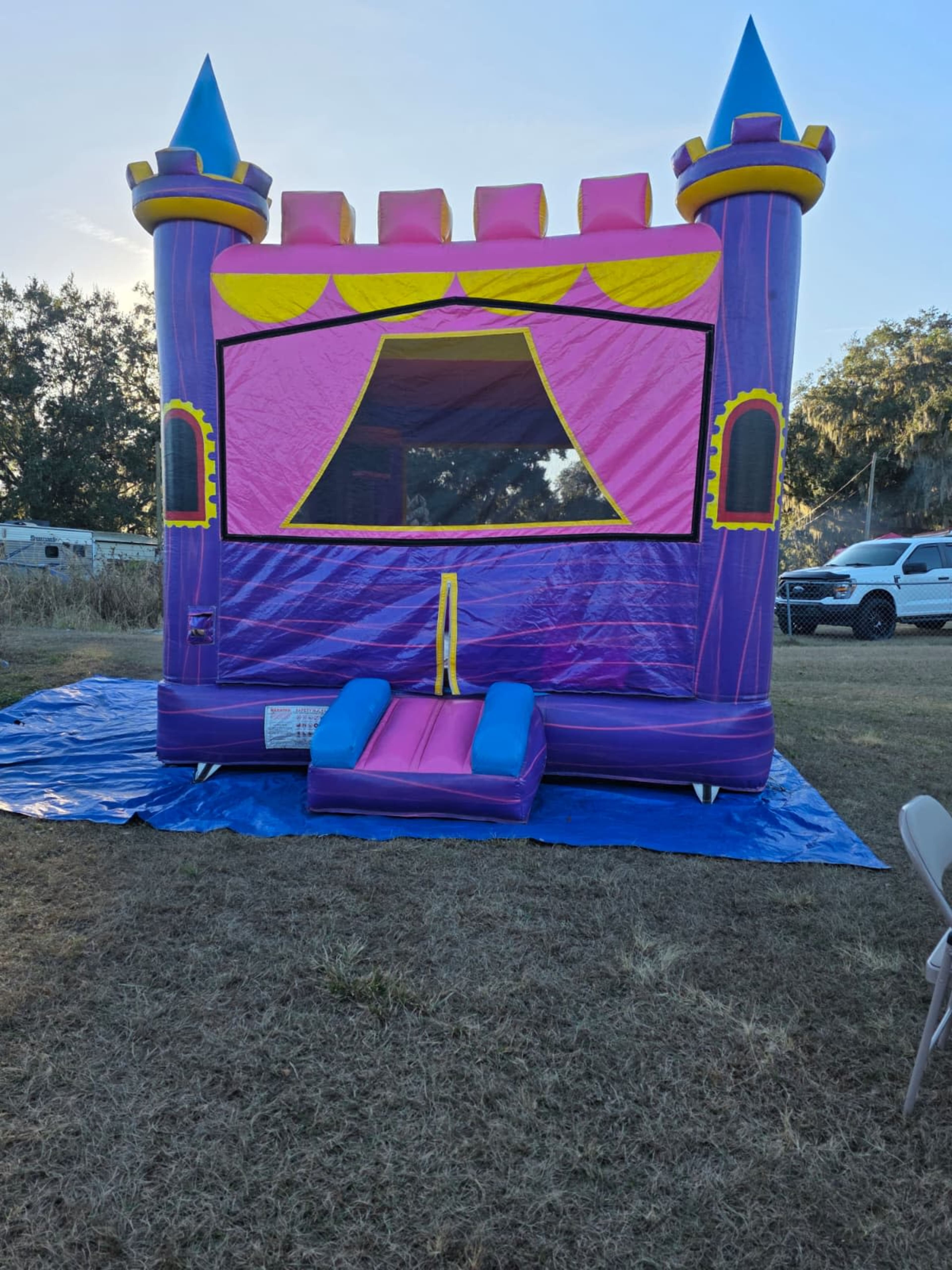A purple and pink inflatable bounce house designed like a castle sits on a blue tarp in a grassy area.