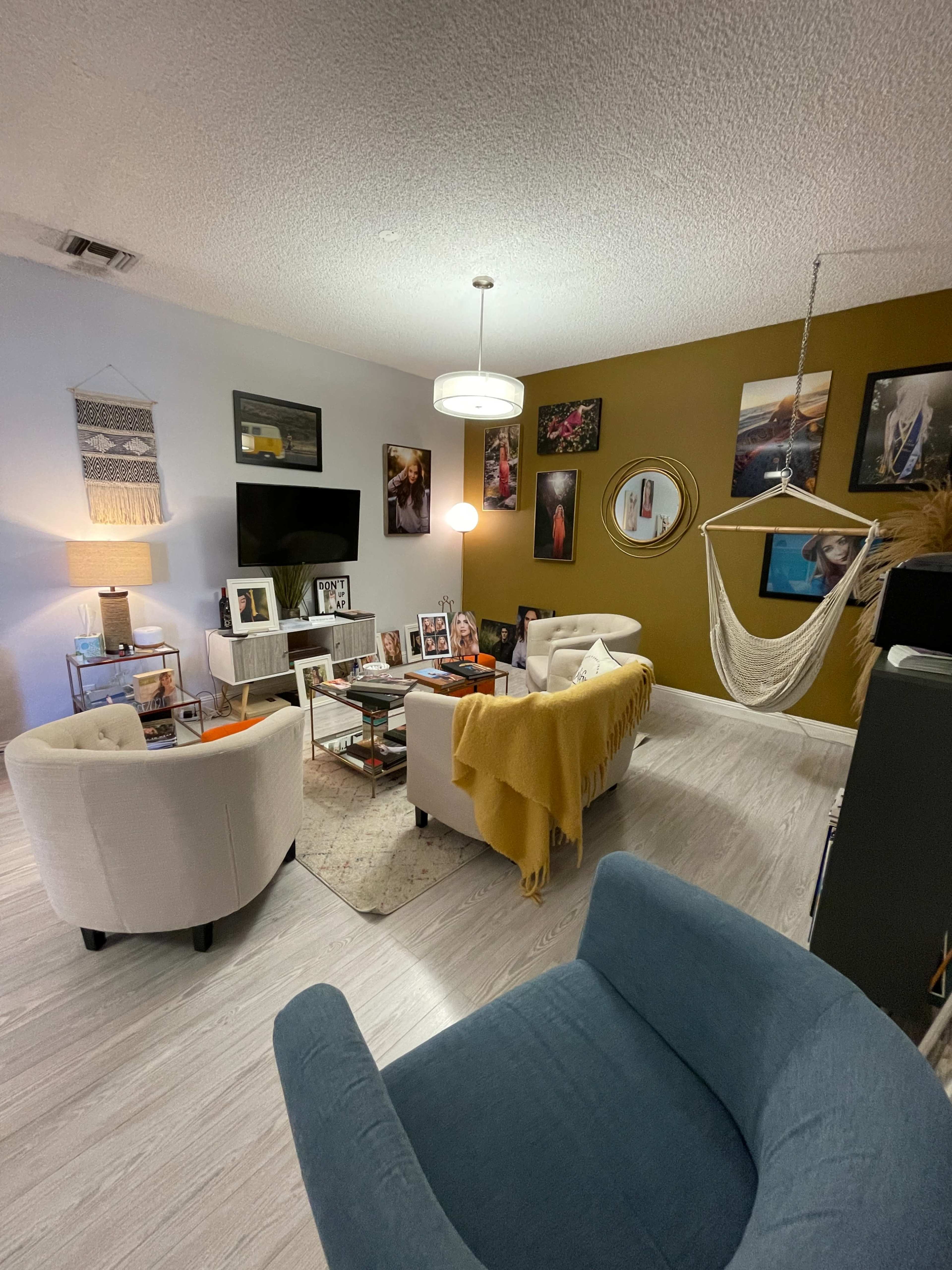 A living room features light-colored walls, a television, comfortable seating, a hammock, and various framed photos on the walls.