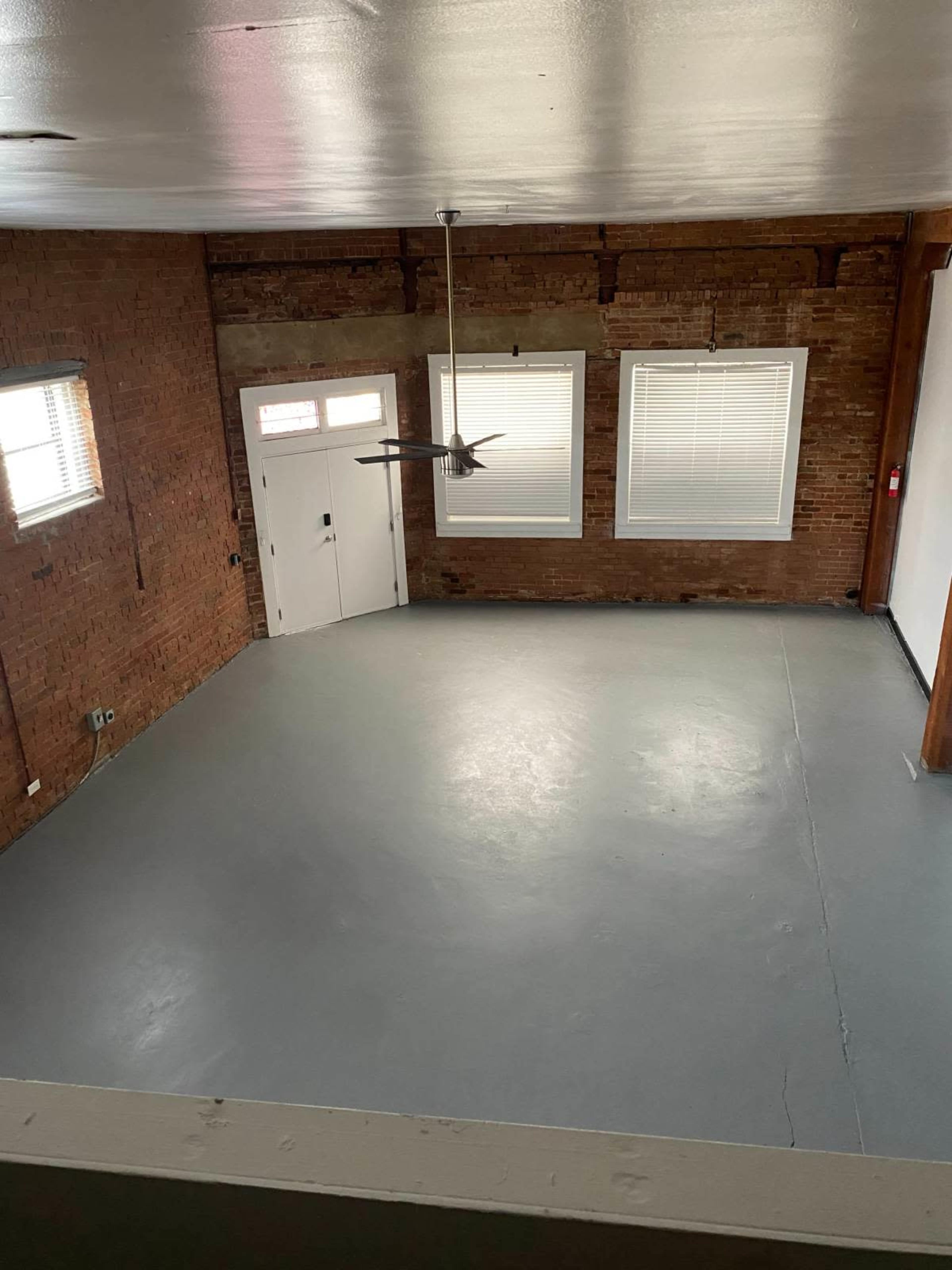 An empty room with exposed brick walls, a concrete floor, and two windows, along with a ceiling fan.