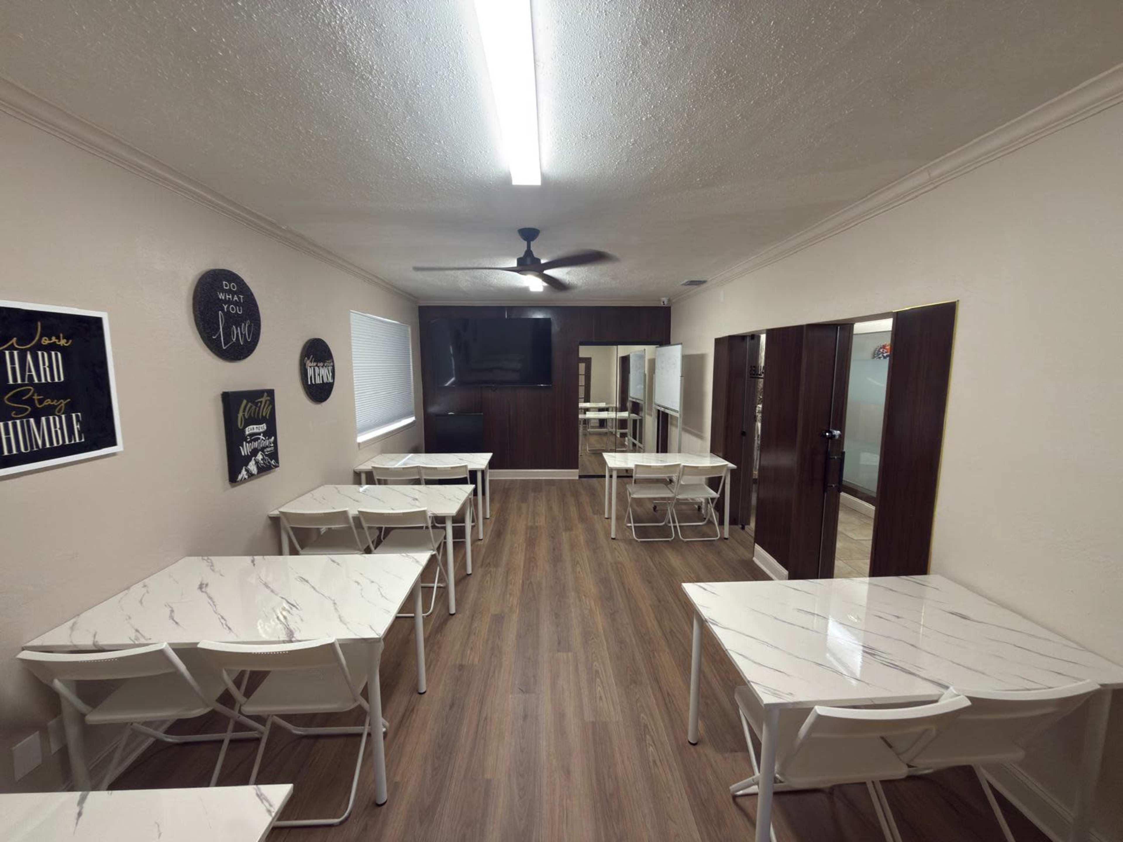 The image shows a clean, spacious room with several white tables and chairs, a ceiling fan, and a wall-mounted television.