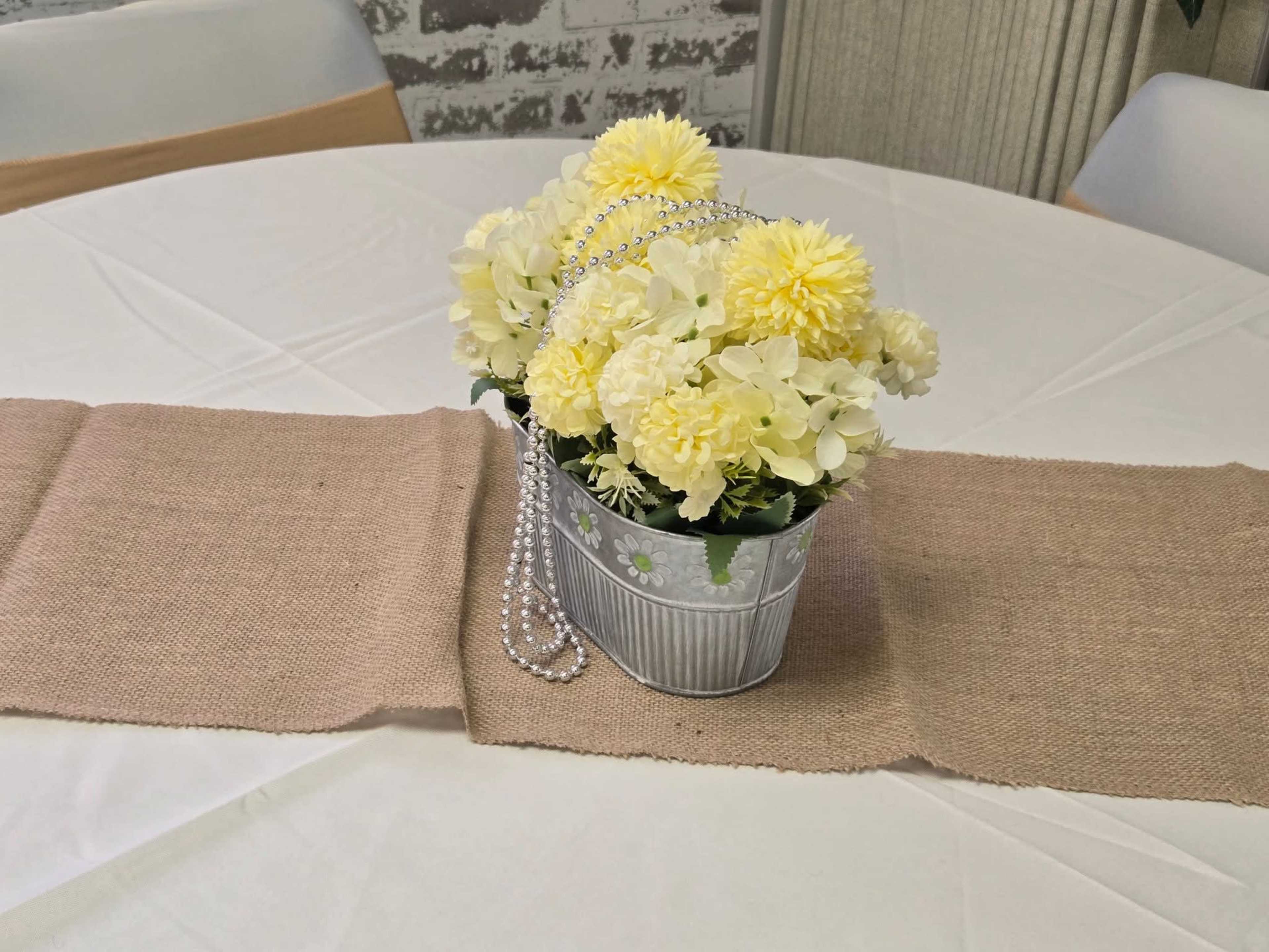 A silver bucket filled with yellow flowers is placed on a table covered with a beige runner.