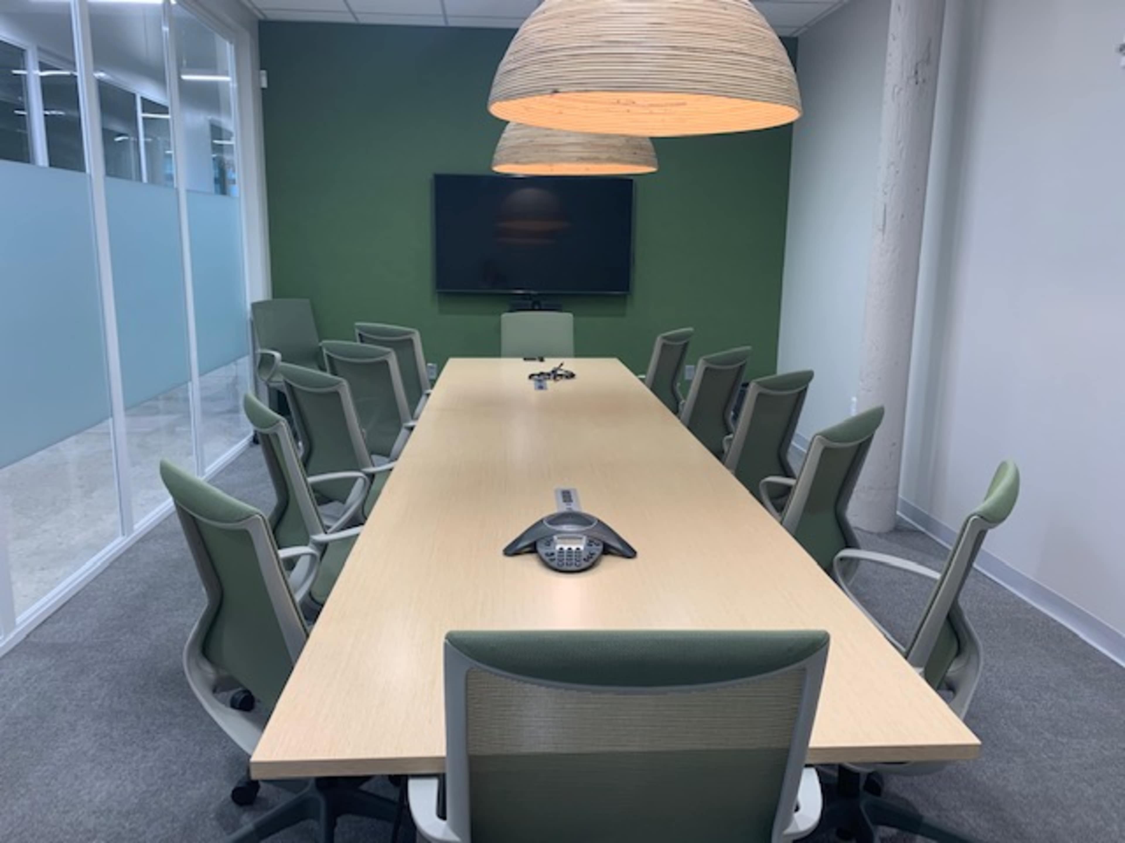 A large conference room features a long wooden table surrounded by green chairs, with two pendant lights hanging above and a wall-mounted screen at the front.