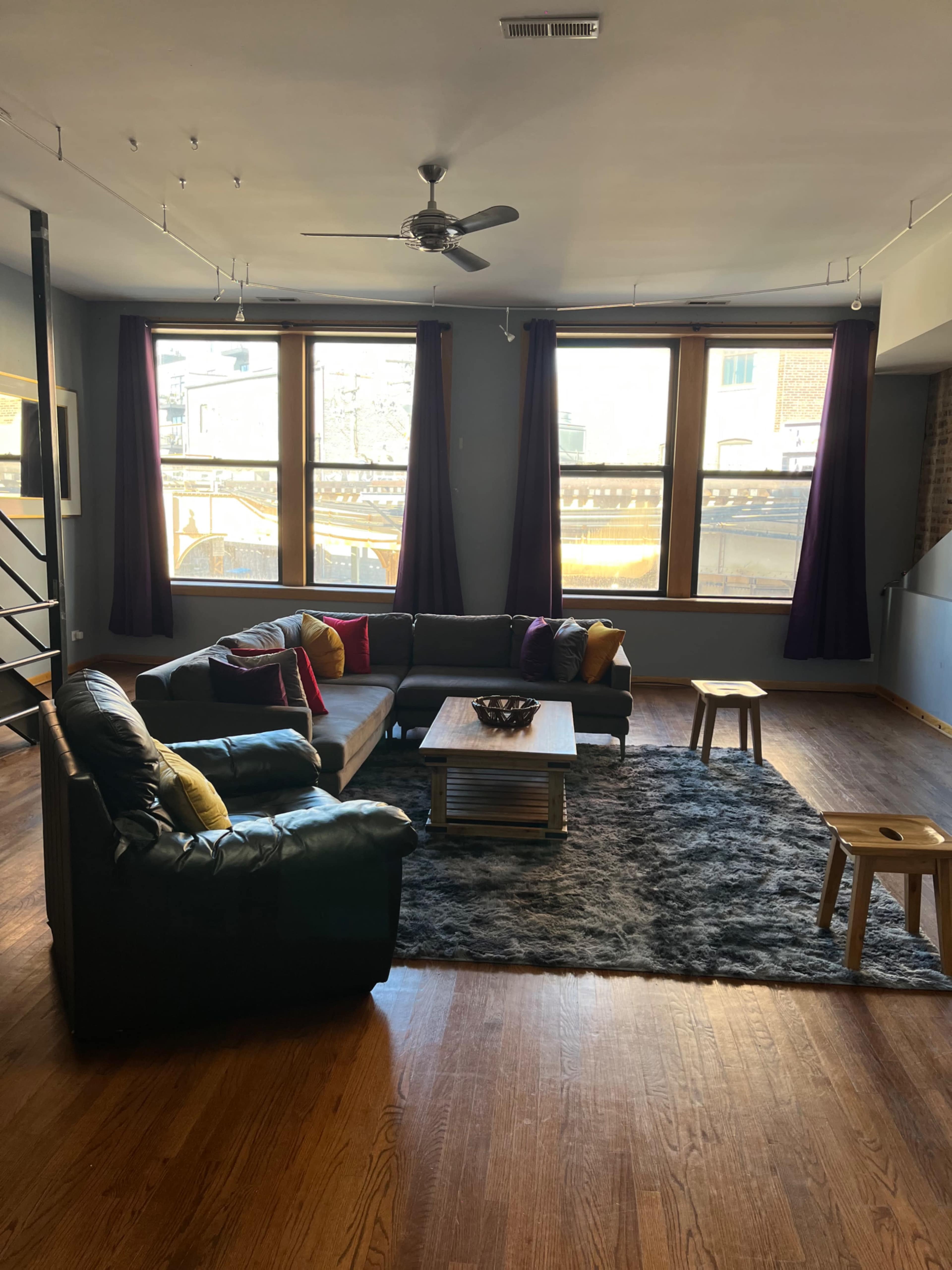 The image shows a spacious living room featuring a black leather sectional sofa, a wooden coffee table on a gray rug, and large windows with dark curtains.