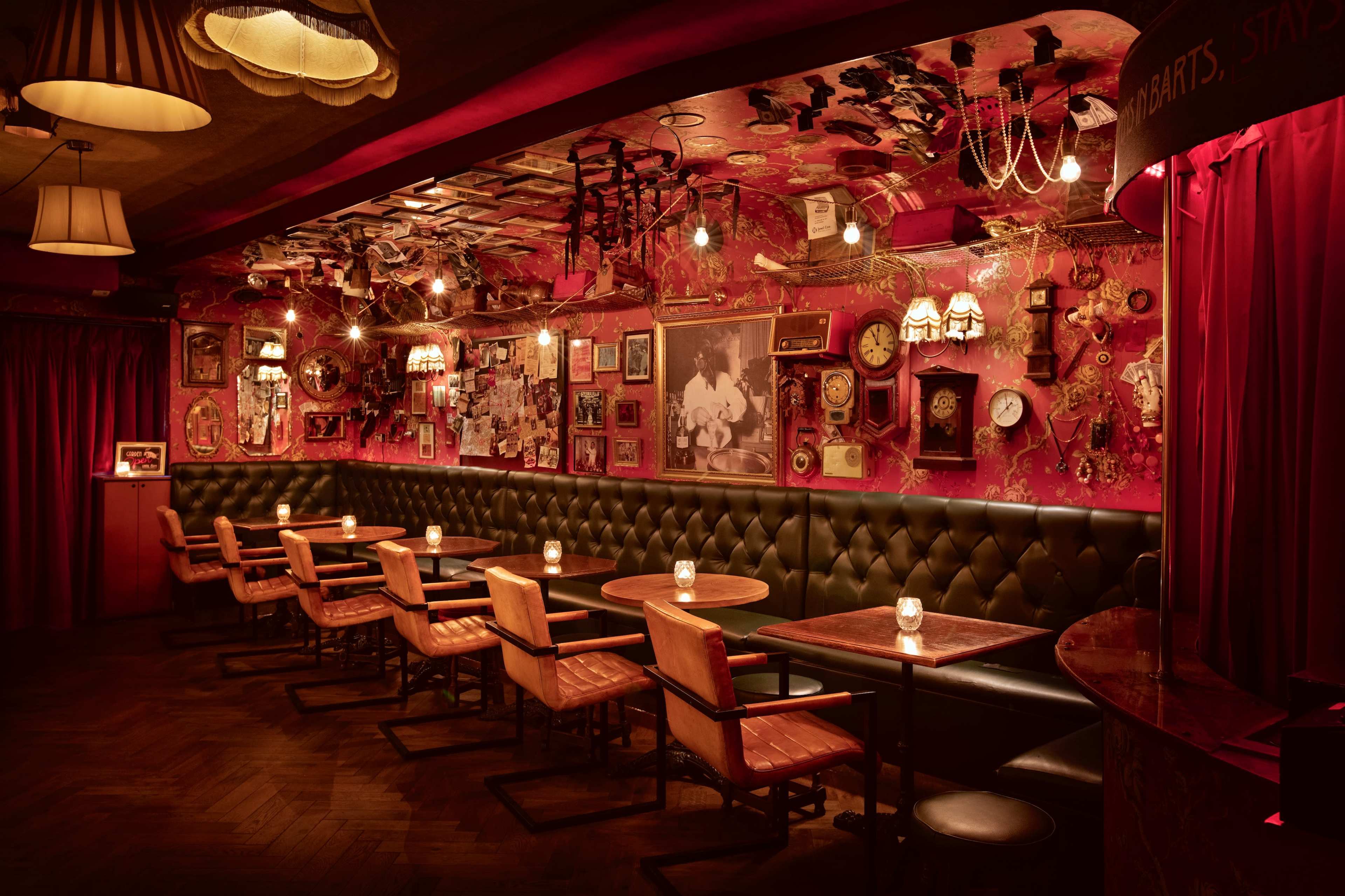 The interior of a warmly-lit bar features dark red walls adorned with vintage decor and small tables arranged along a leather-upholstered banquette.