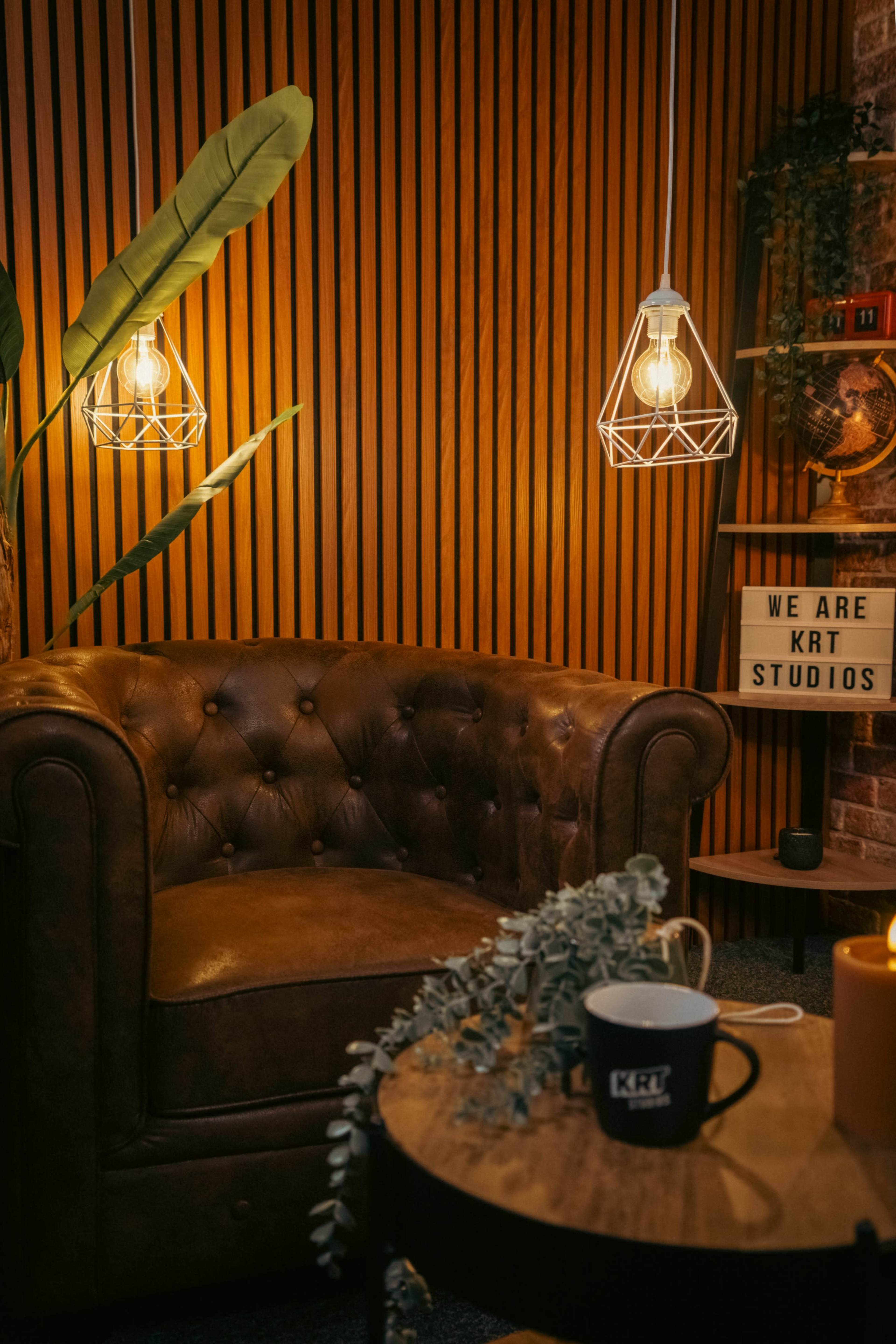 The image shows a cozy interior featuring a brown leather tufted couch, pendant lights, and a decorative plant against a wooden panel wall.