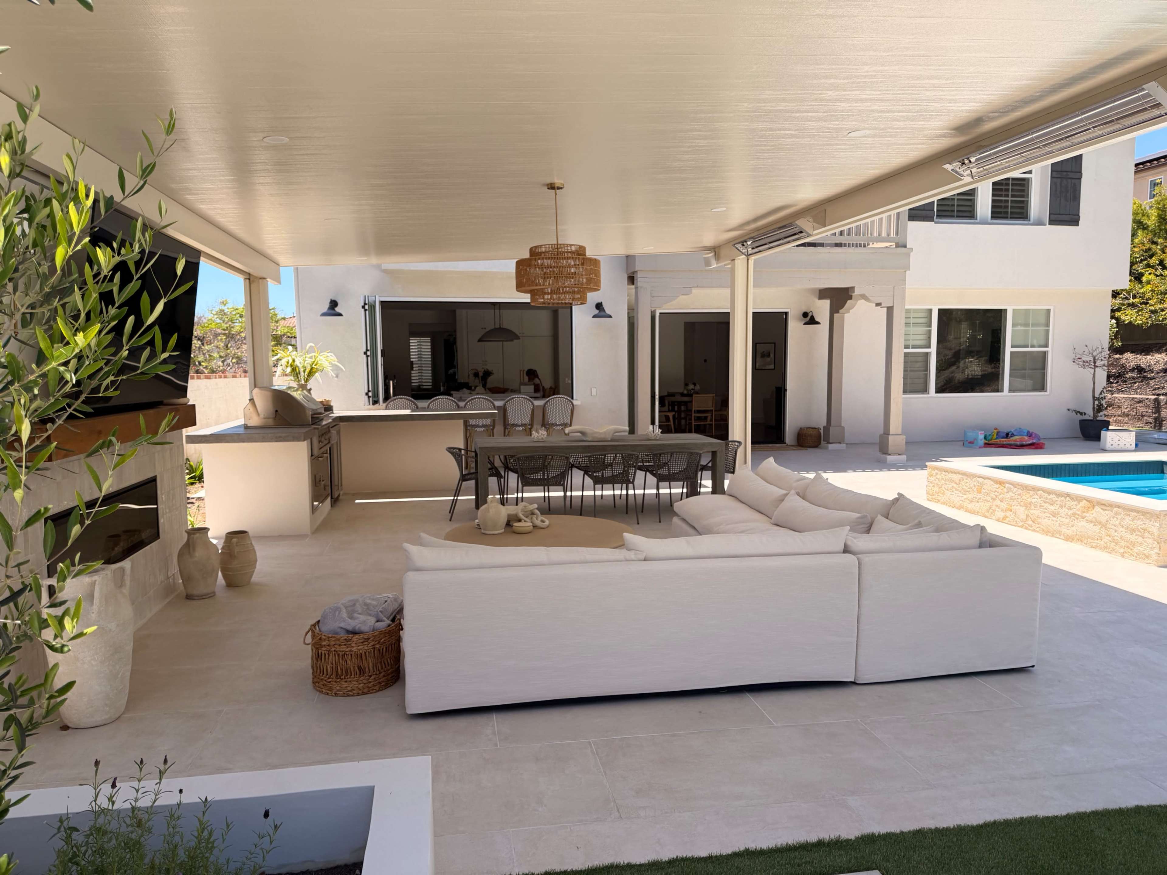 The image shows an outdoor living area featuring a large white couch, a dining table, and a pool, with a modern kitchen in the background.