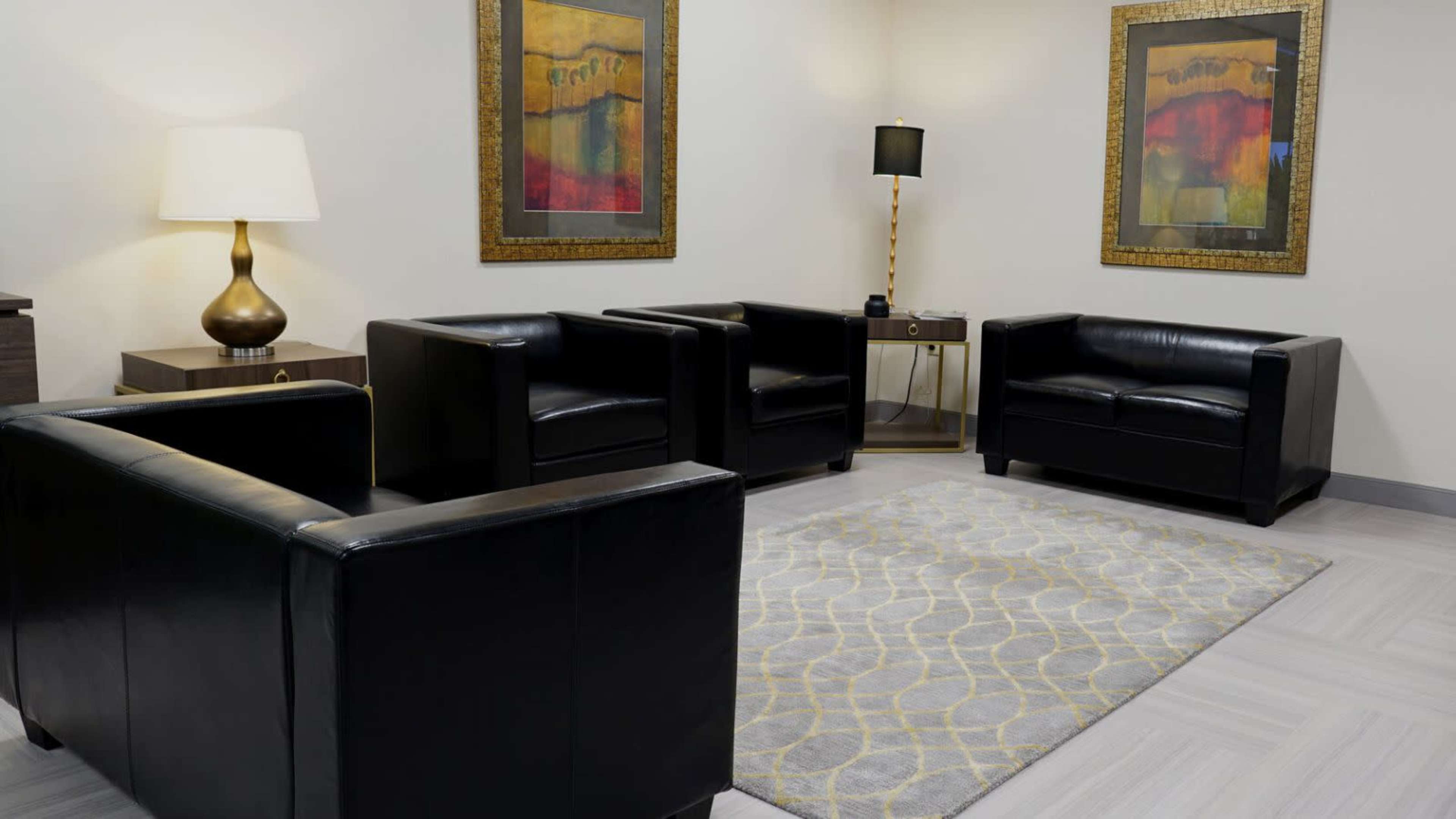 The image features a waiting room with two black leather chairs, a rug with a geometric pattern, and framed artwork on the walls, accompanied by a lamp and a small table.