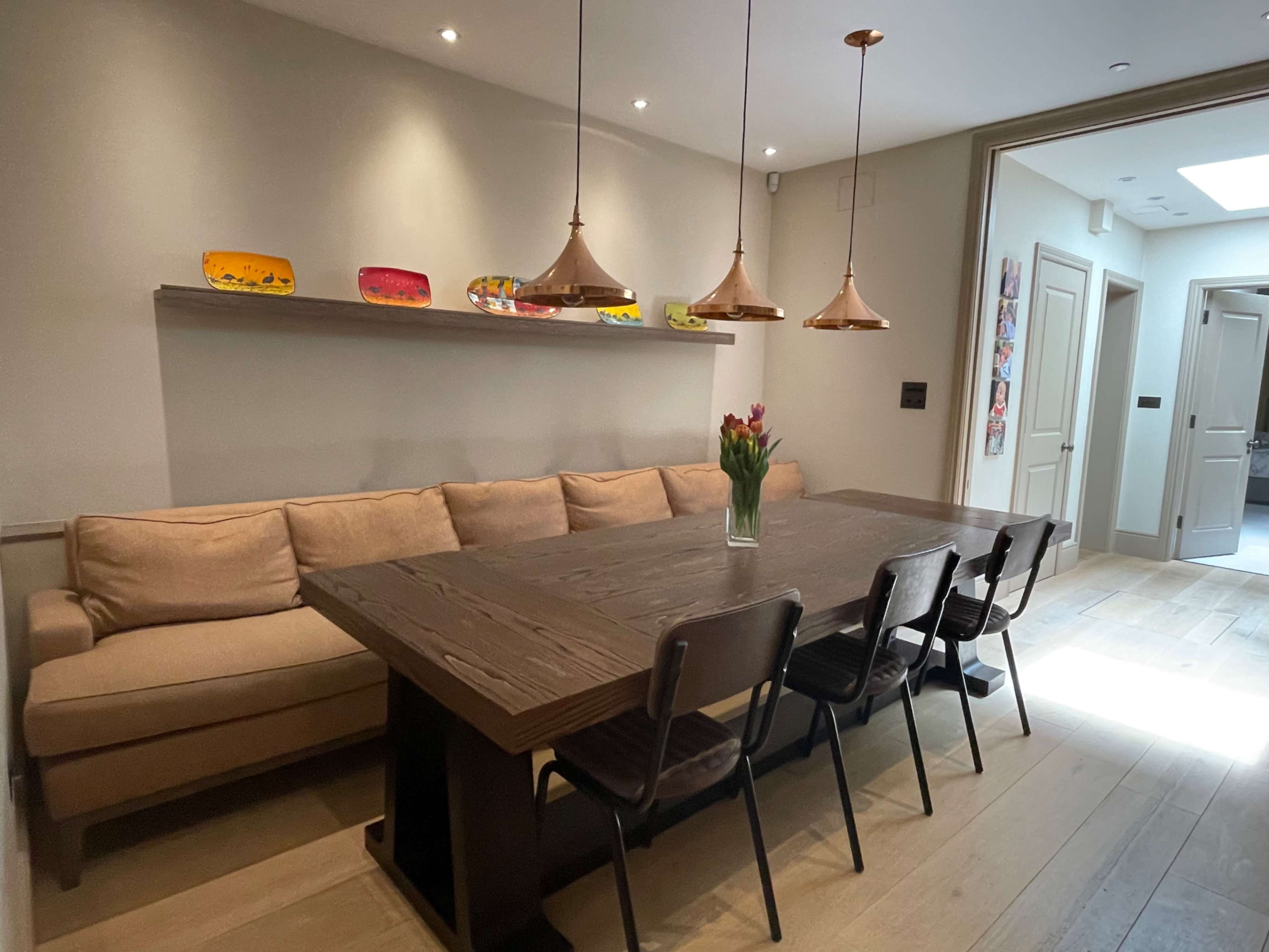 A long wooden dining table surrounded by black chairs is set against a wall with a shelf displaying colorful decorative plates, with a beige sofa alongside it.