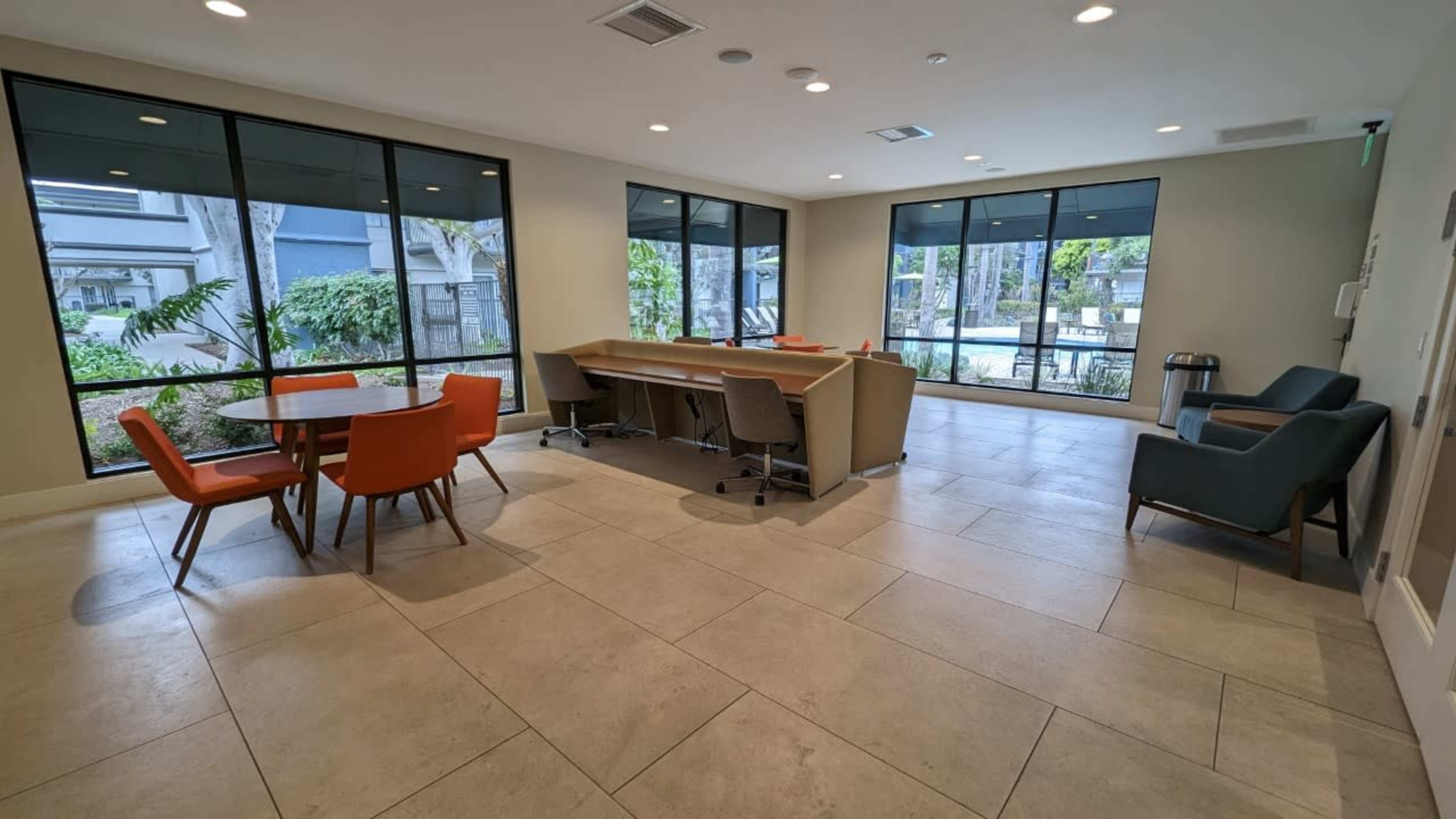 A modern common area with large windows, featuring a dining table with orange chairs, a work desk, and a couple of chairs in a neutral-toned space.