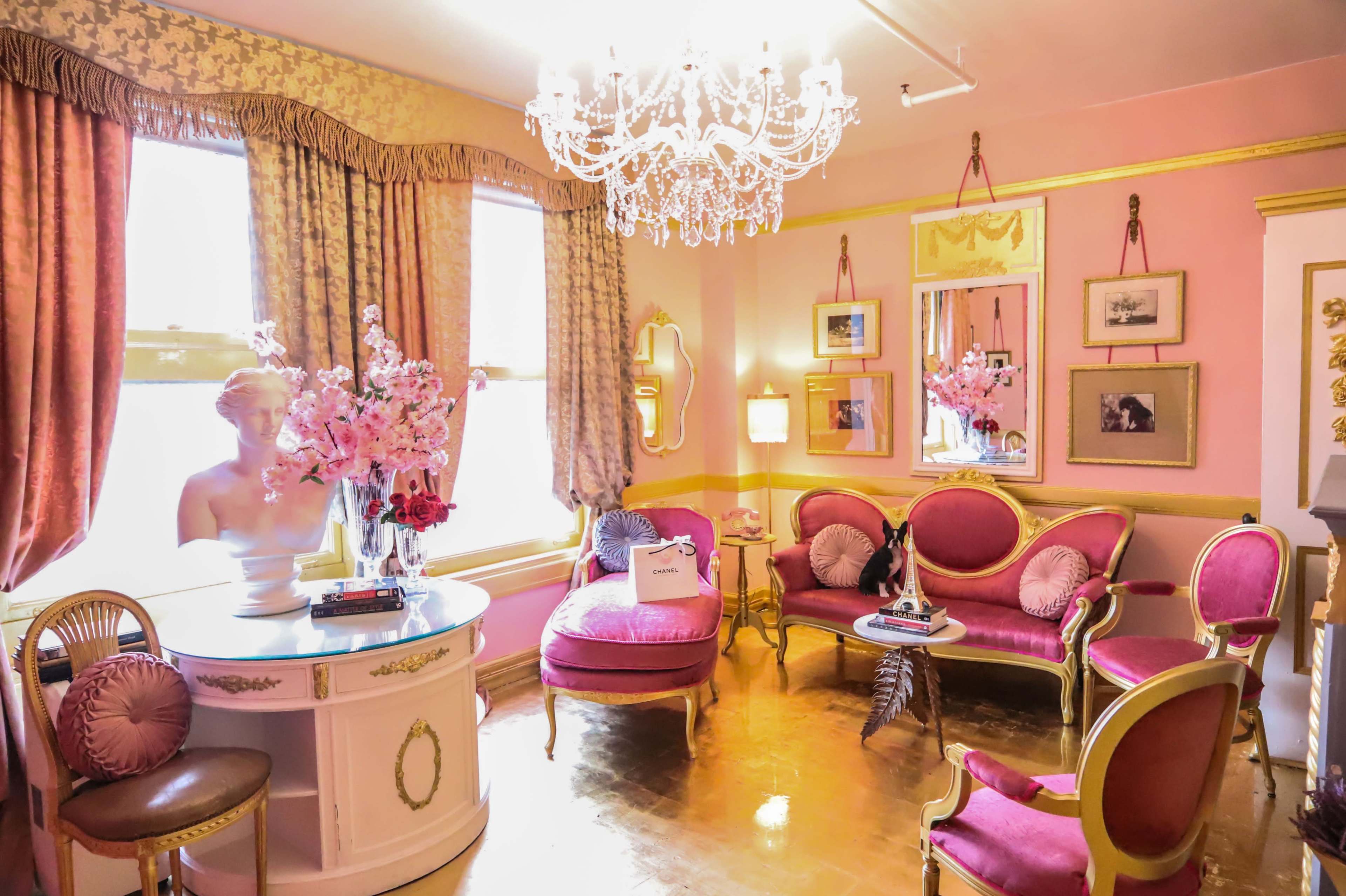 A pink-themed living room featuring a chandelier, a bust of a woman, a round table, and upholstered seating adorned with floral arrangements.