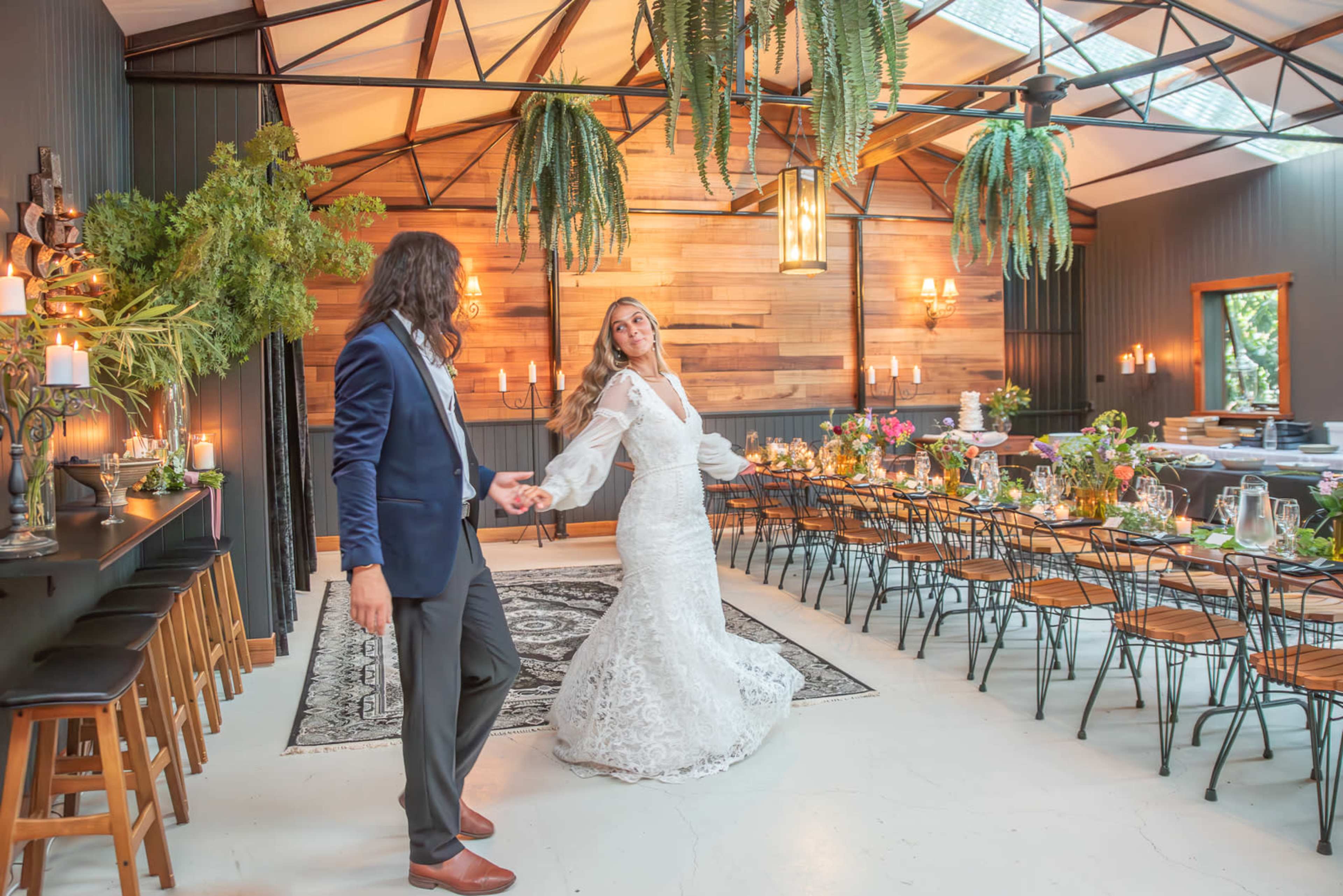 A couple dances in a beautifully decorated indoor venue with wooden walls, greenery, and a long dining table set for a celebration.