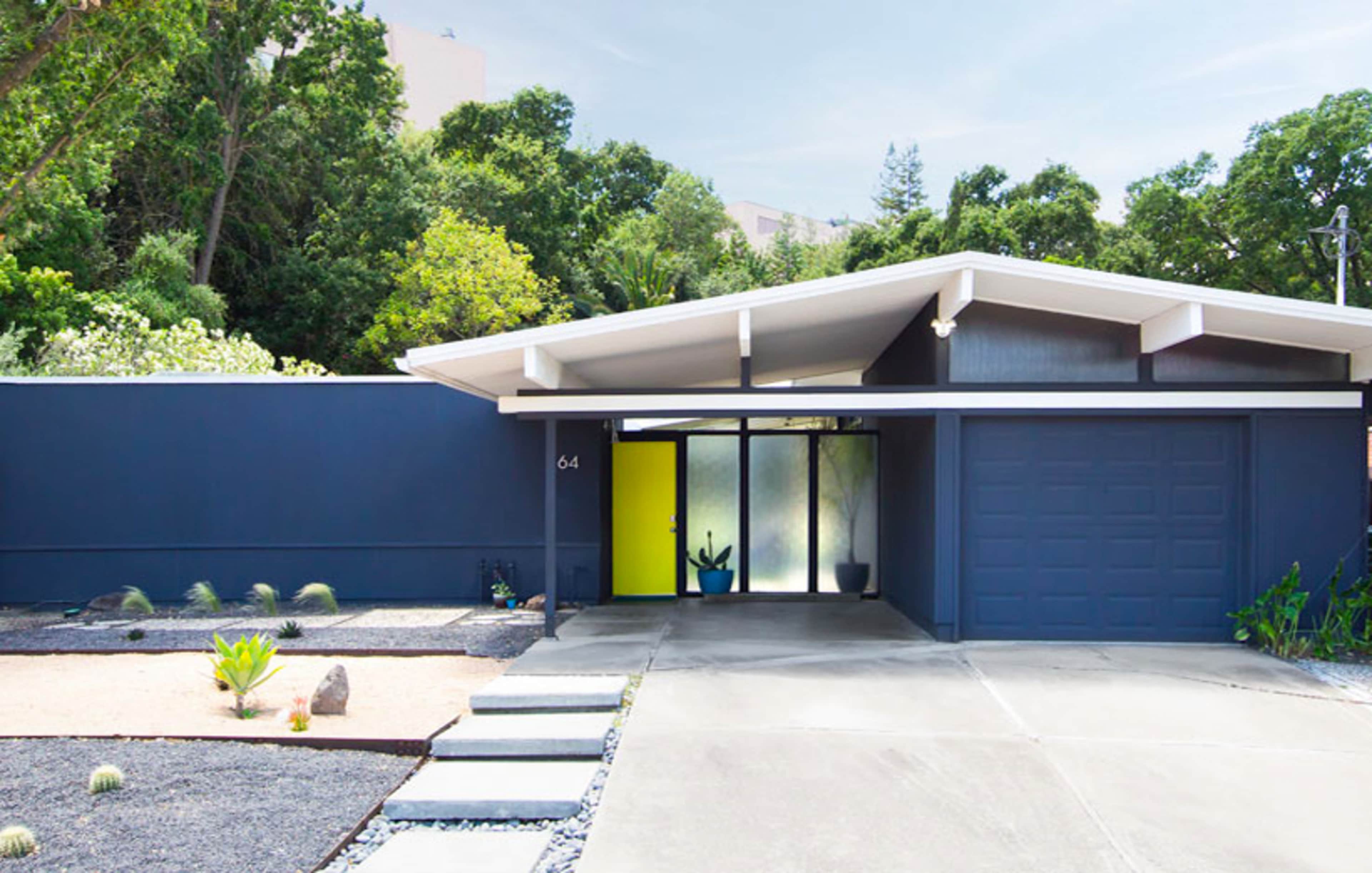 A modern house with a flat roof, a large glass entrance featuring a bright yellow door, and a landscaped front yard.