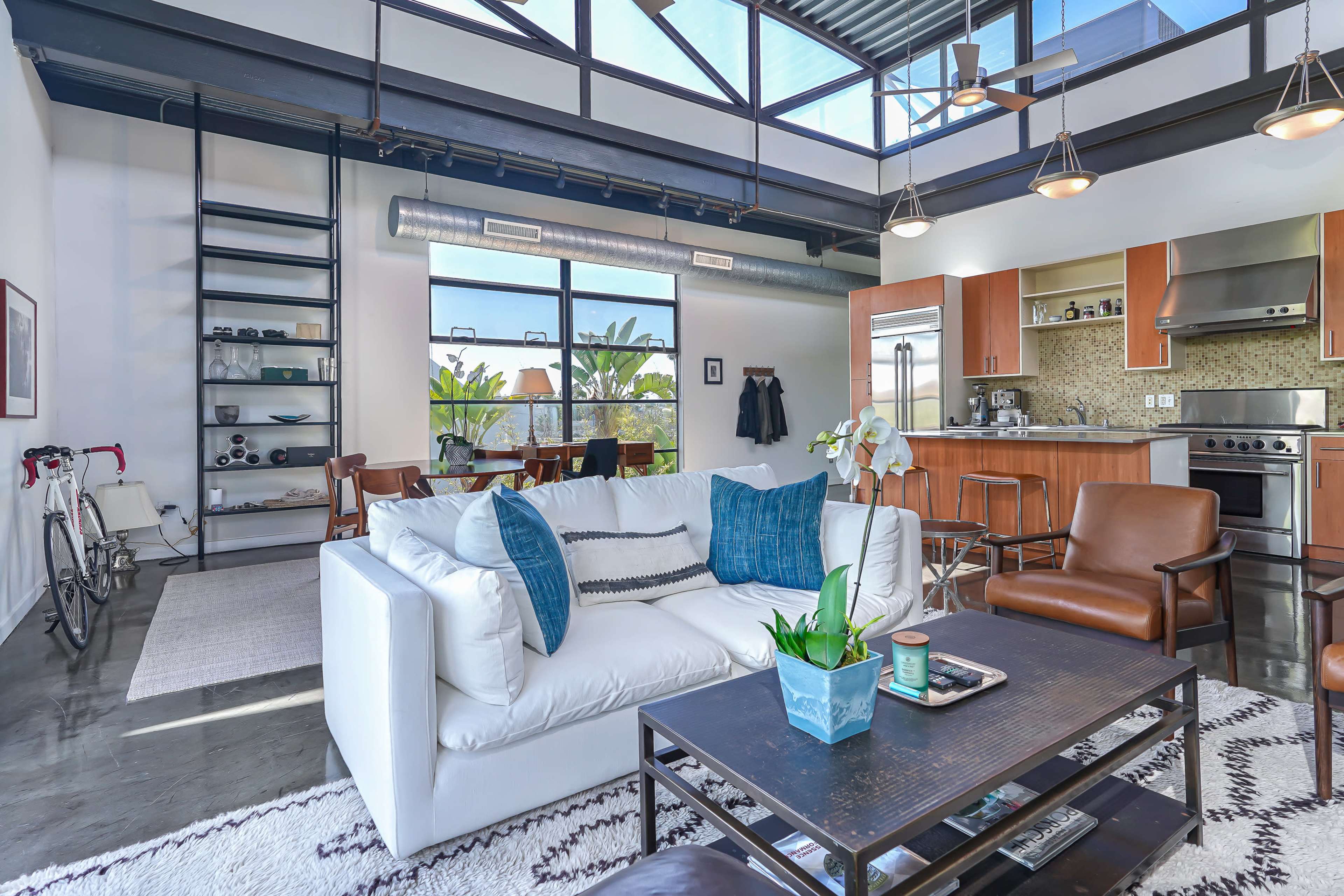 The image shows a modern loft-style living space with a white sofa, a wooden coffee table, and a kitchen area visible in the background.