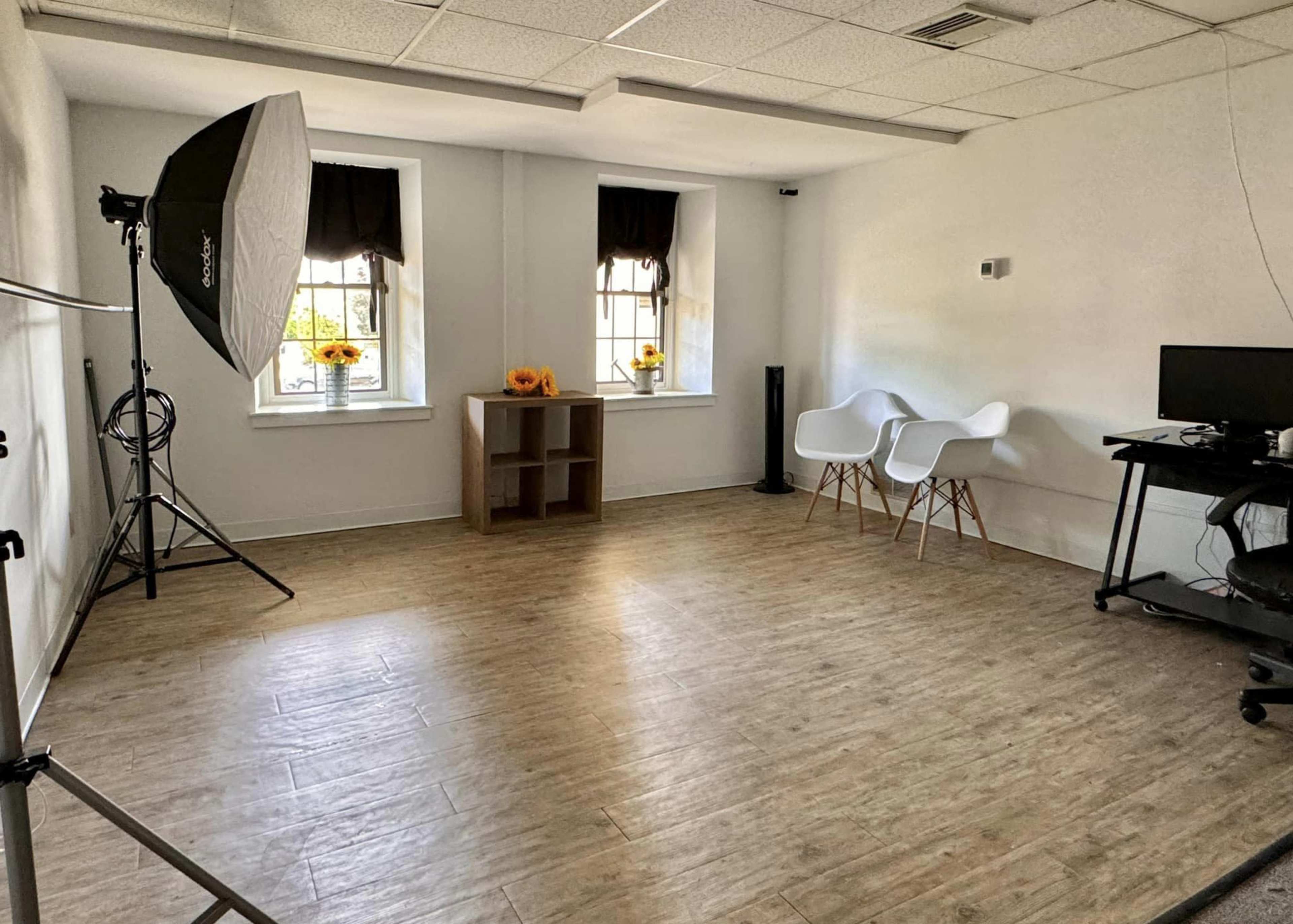 The image shows a sparsely furnished studio space with two white chairs, a small shelf, a computer workstation, and lighting equipment.
