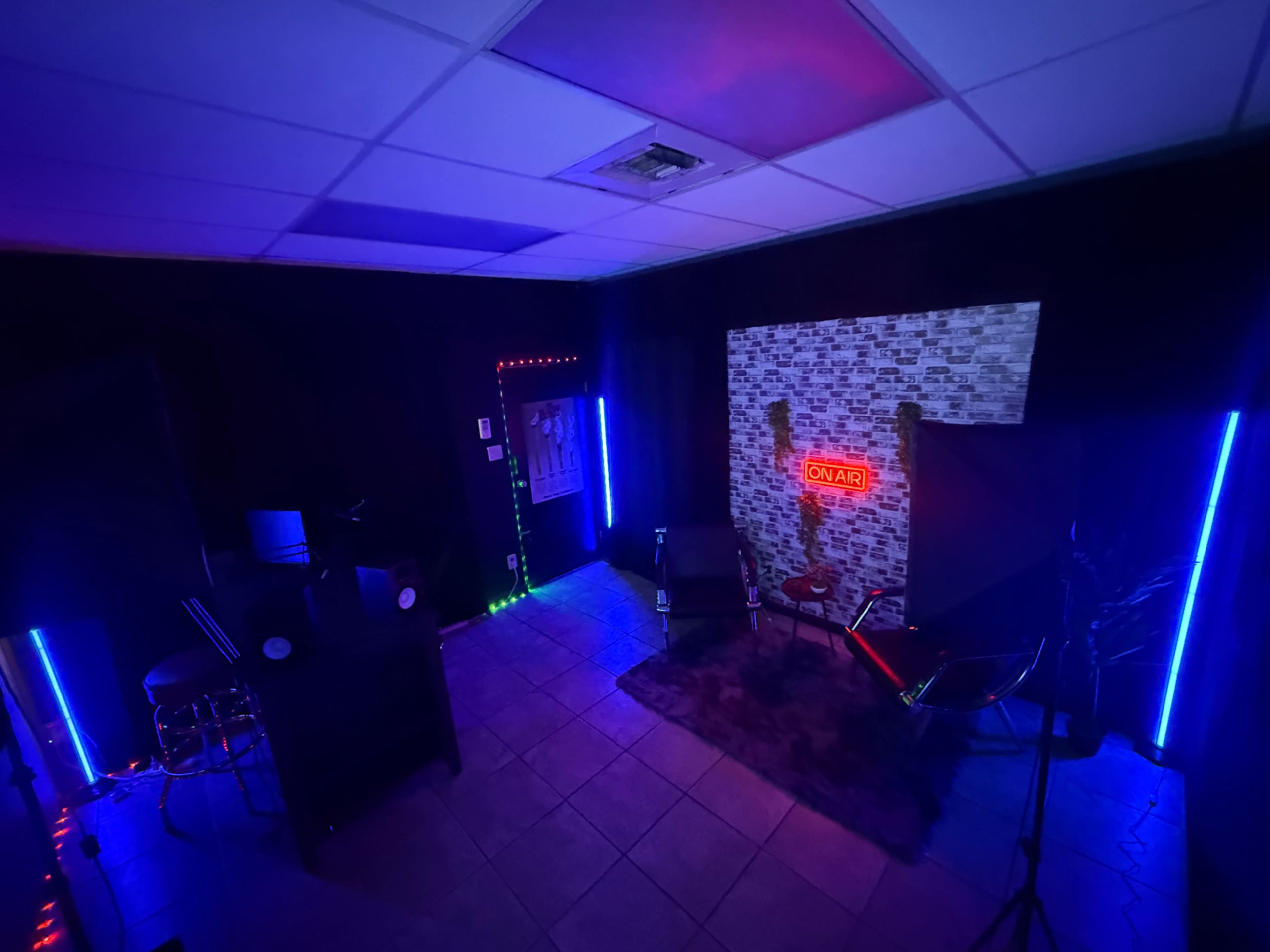 The image shows a dimly lit studio with neon lights, a brick wall featuring an "ON AIR" sign, and various seating arrangements.