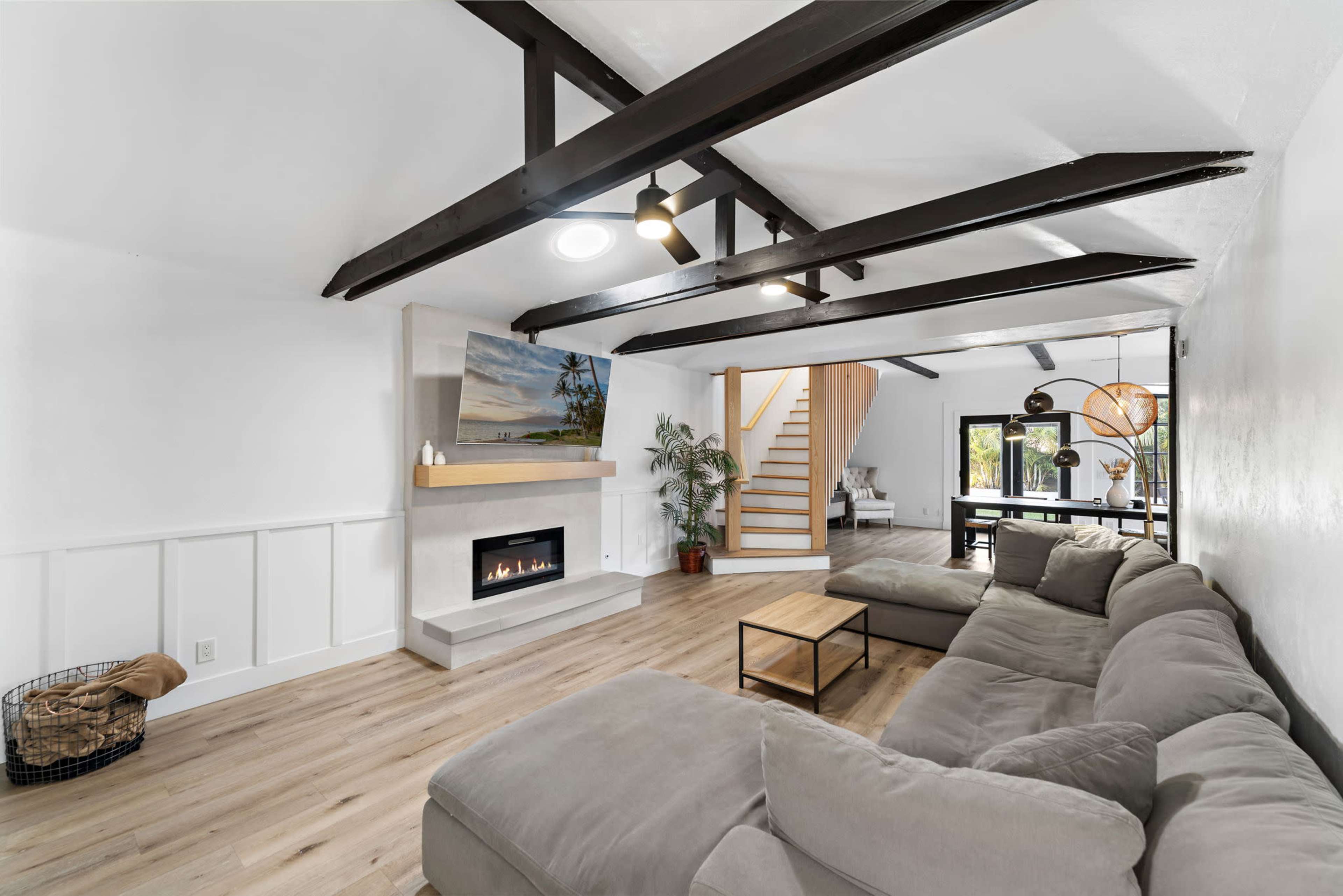 The image shows a modern living room featuring a large sectional sofa, a sleek fireplace, and a staircase leading to an upper level, with wood beams overhead and a neutral color palette.