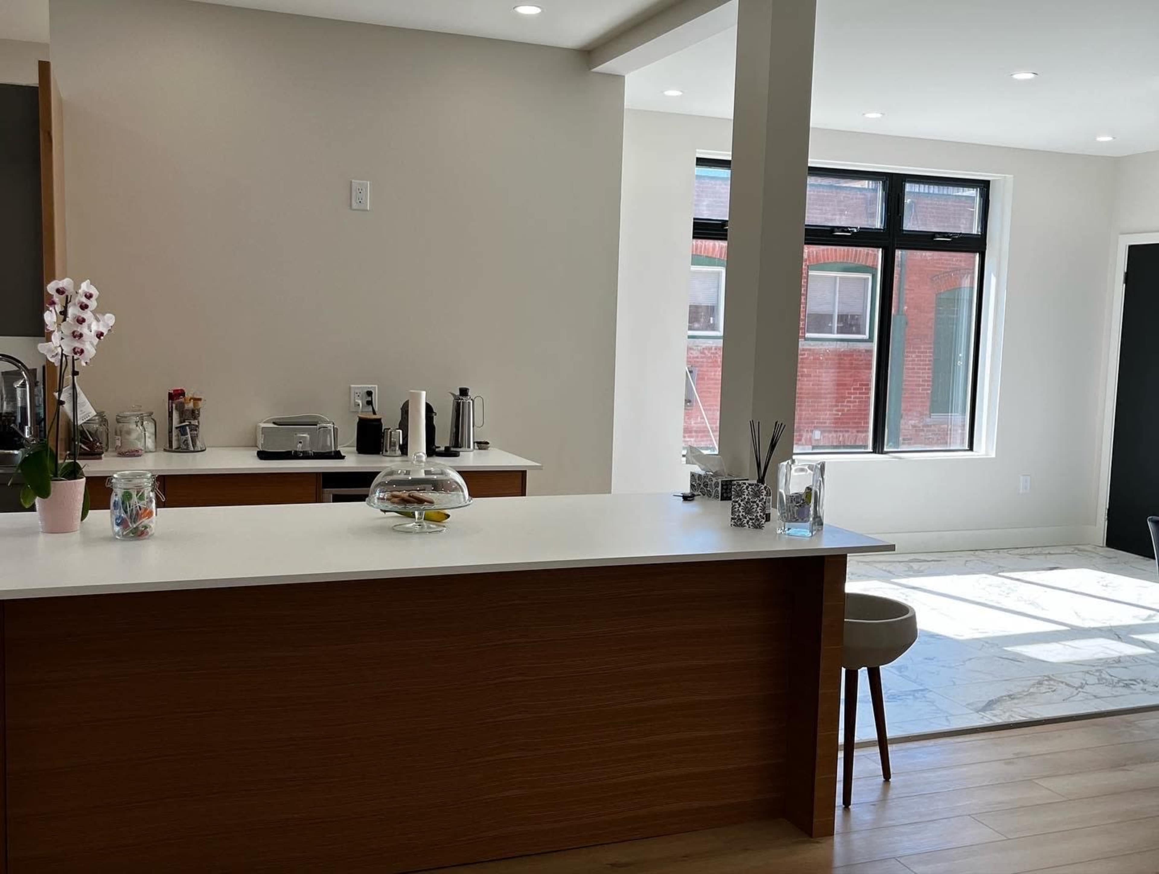 A modern kitchen features a central island with a glass cloche and a seating area, adjacent to a brightly lit dining space with large windows.
