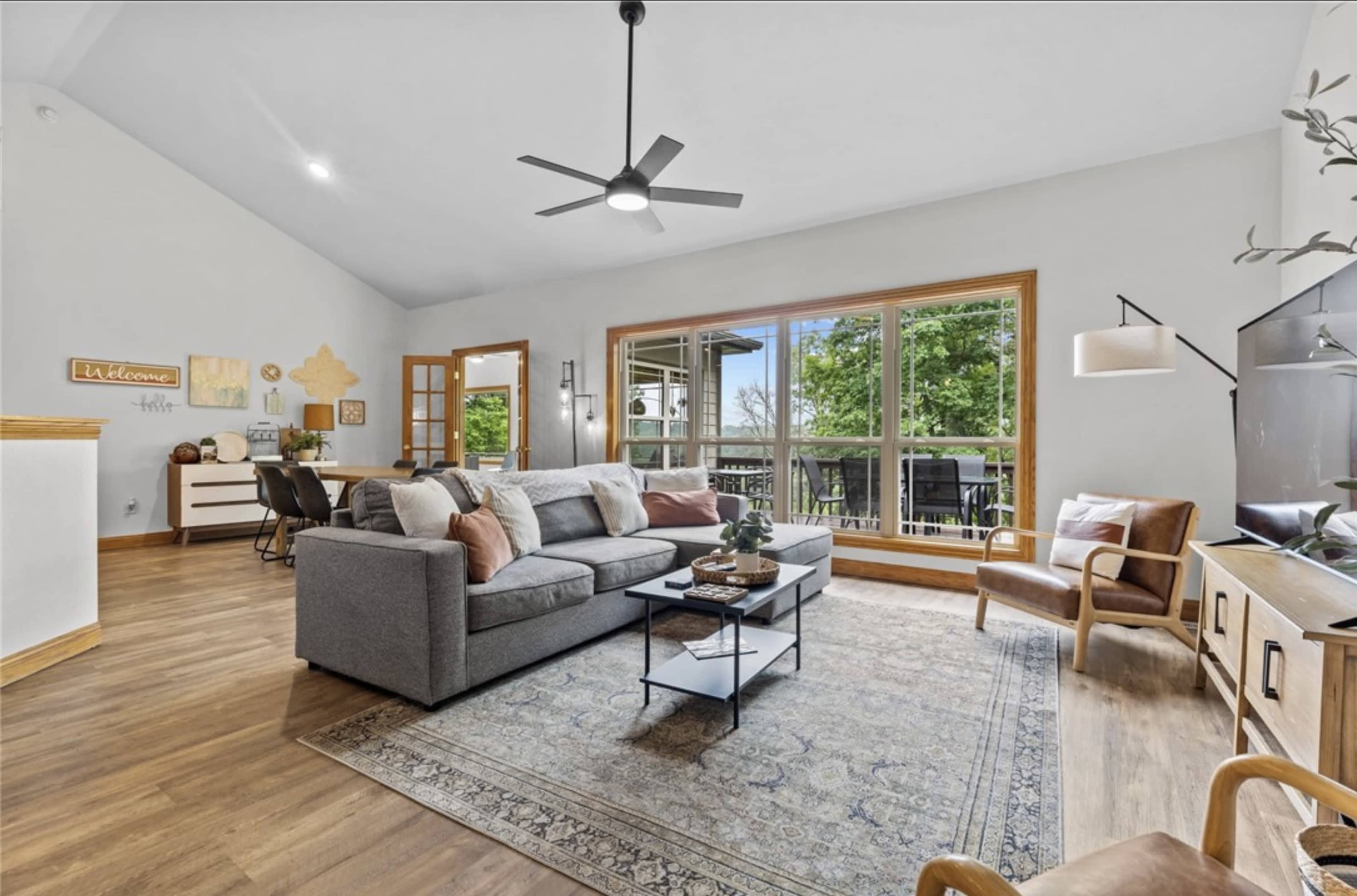 The living room features a gray sectional sofa, a coffee table, and large windows that overlook a balcony area.