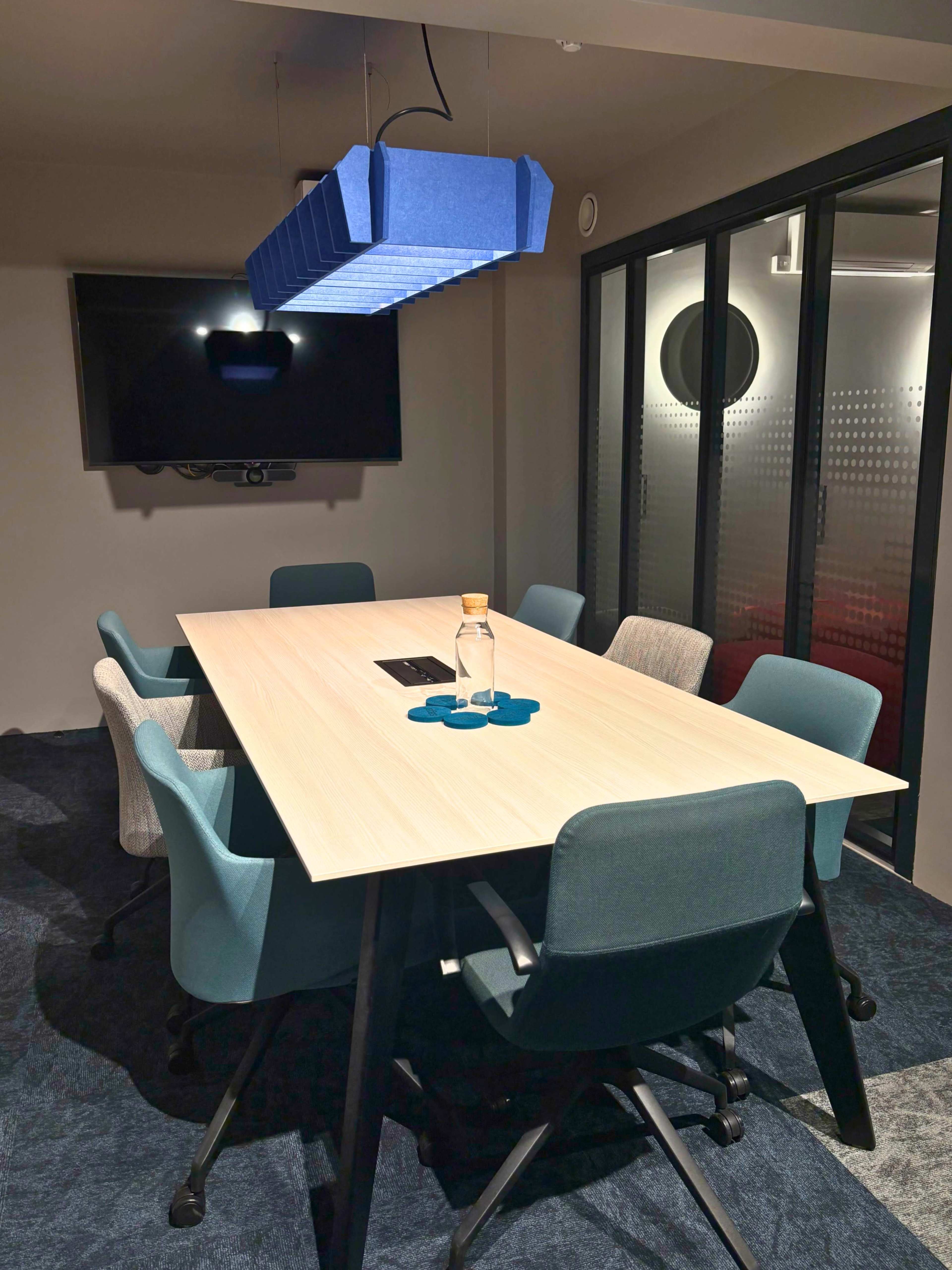 A modern conference room features a long table surrounded by six chairs, with a large screen and a blue acoustic light fixture overhead.