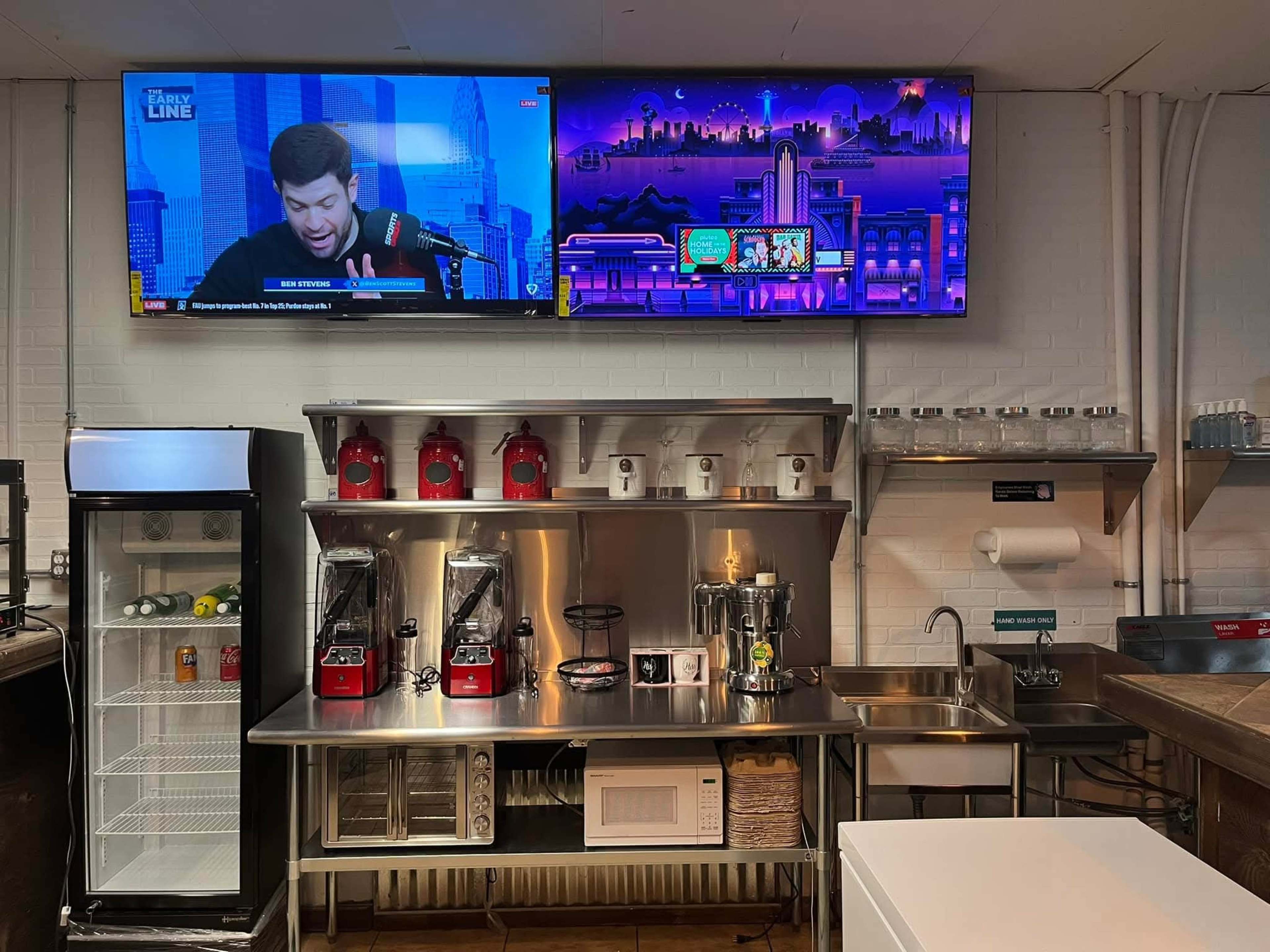 The image shows a cafe interior with an L-shaped counter featuring coffee machines, dispensers, and a wall-mounted TV displaying a program, alongside a refrigerator and glassware on shelves.