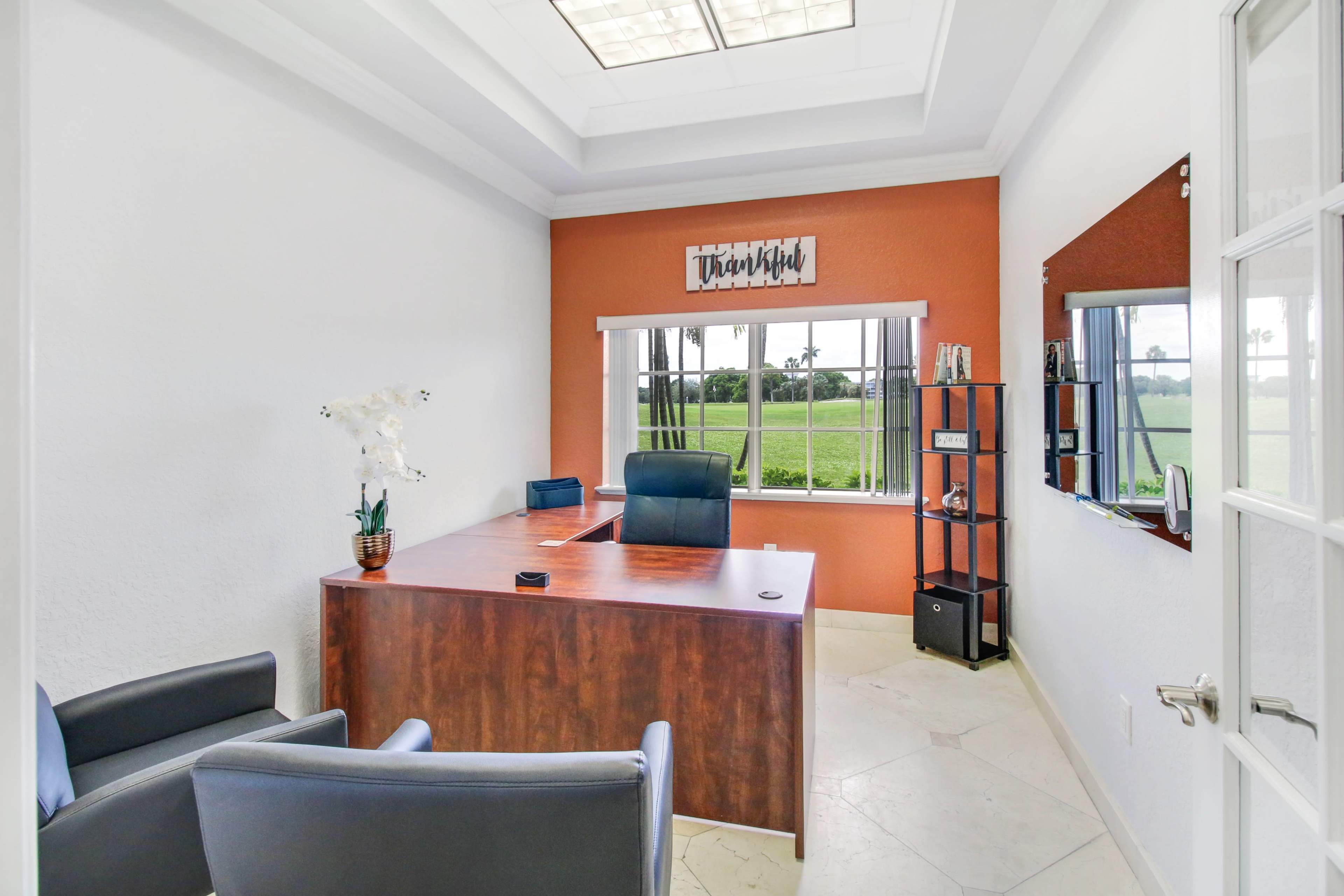 The image shows a small office with a wooden desk, two chairs, a bookshelf, and a large window overlooking a green landscape.