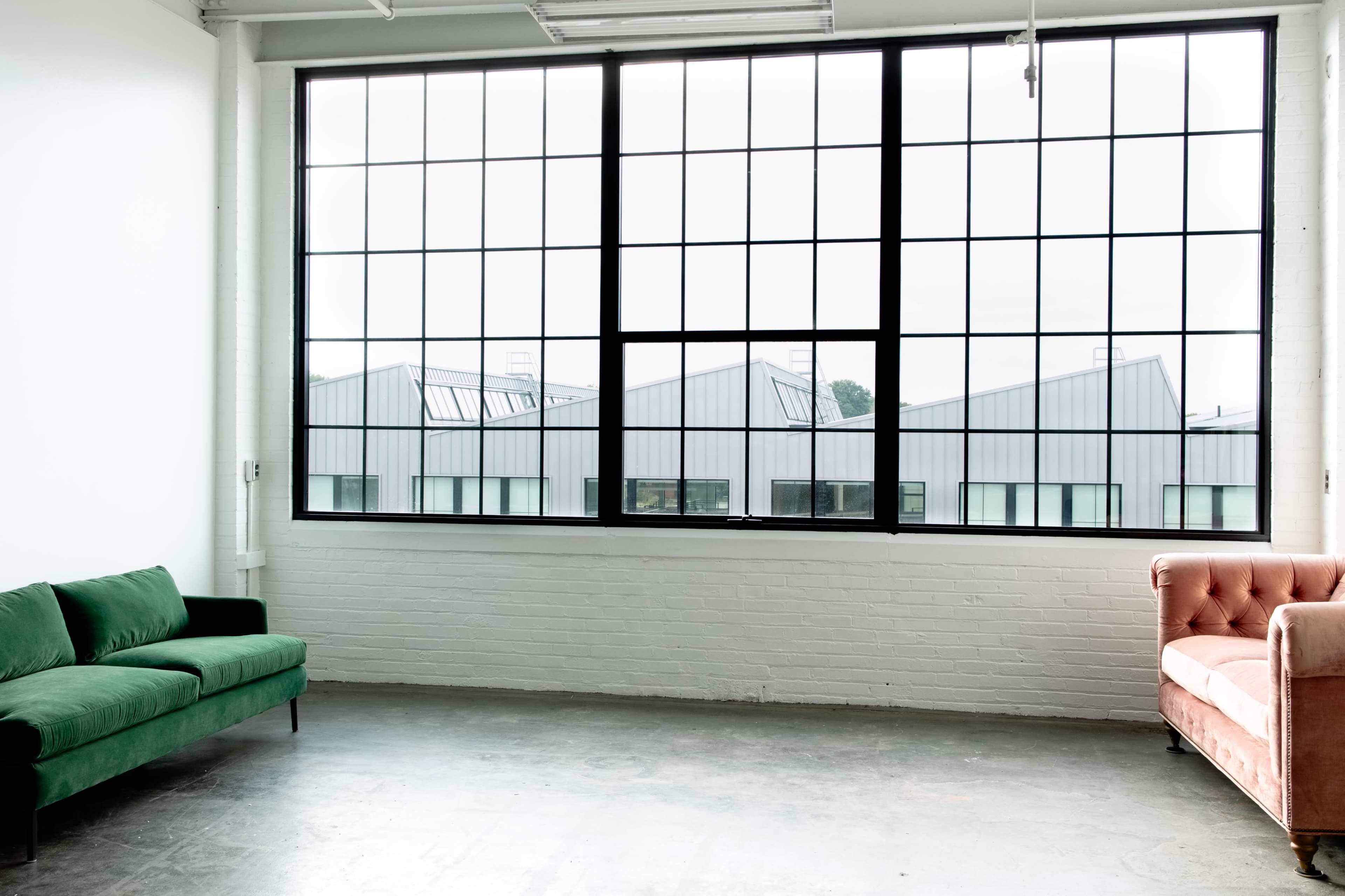 The image shows a spacious room with large windows overlooking industrial-style buildings, featuring a green sofa and a pink armchair against the white walls.