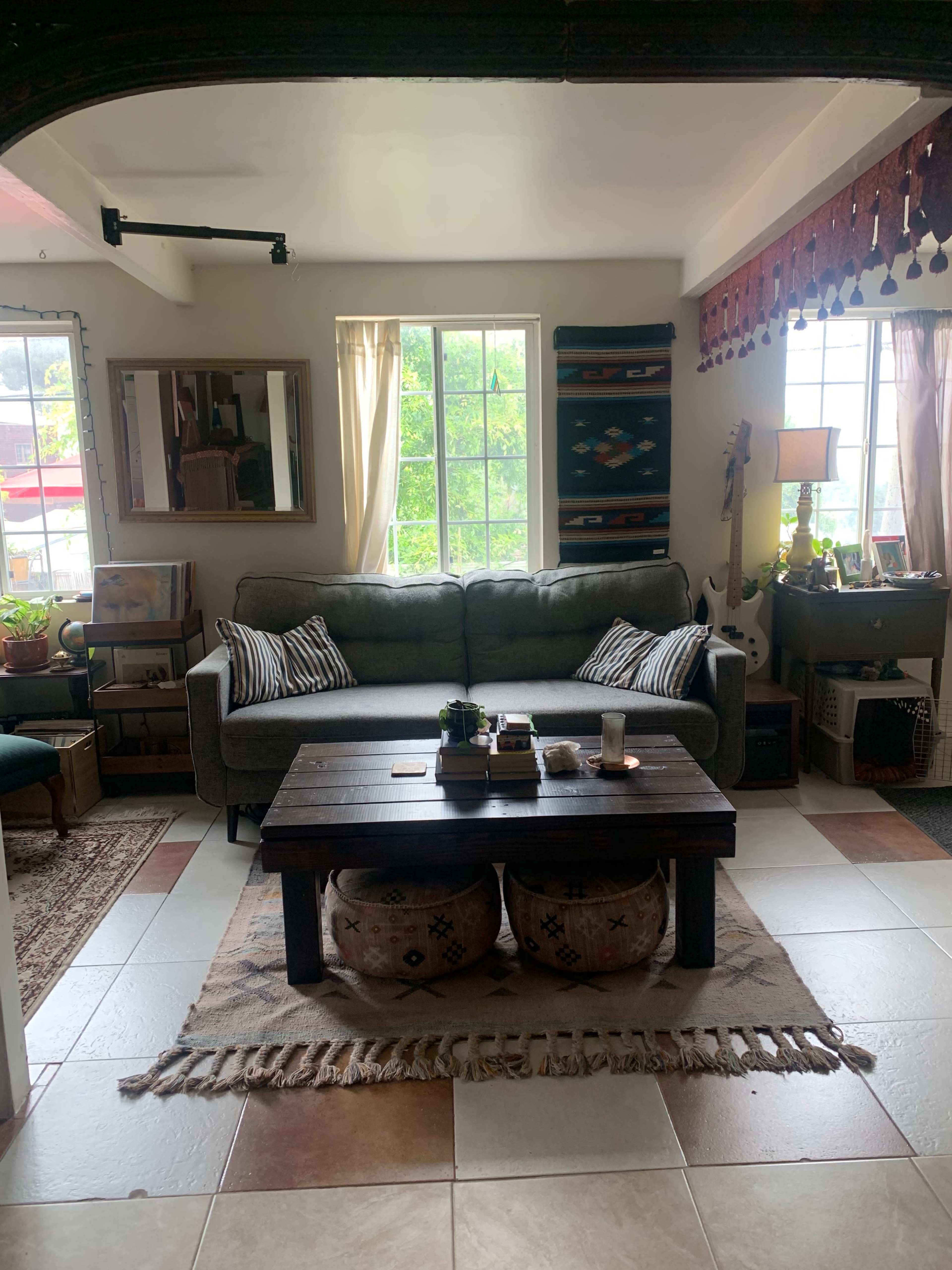 A cozy living room featuring a gray sofa, a wooden coffee table with decorative items, two upholstered stools, and a bright window with plants and artwork around.
