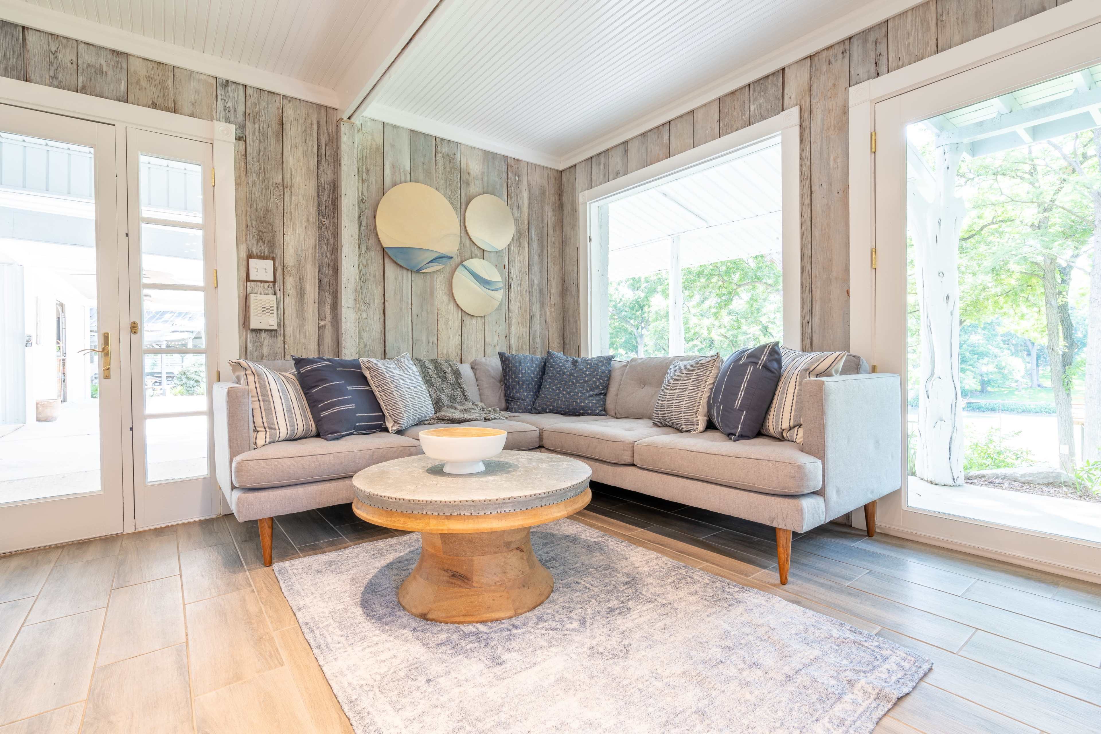 A modern living room features a gray sectional sofa, a round wooden coffee table, and large windows with views of greenery.
