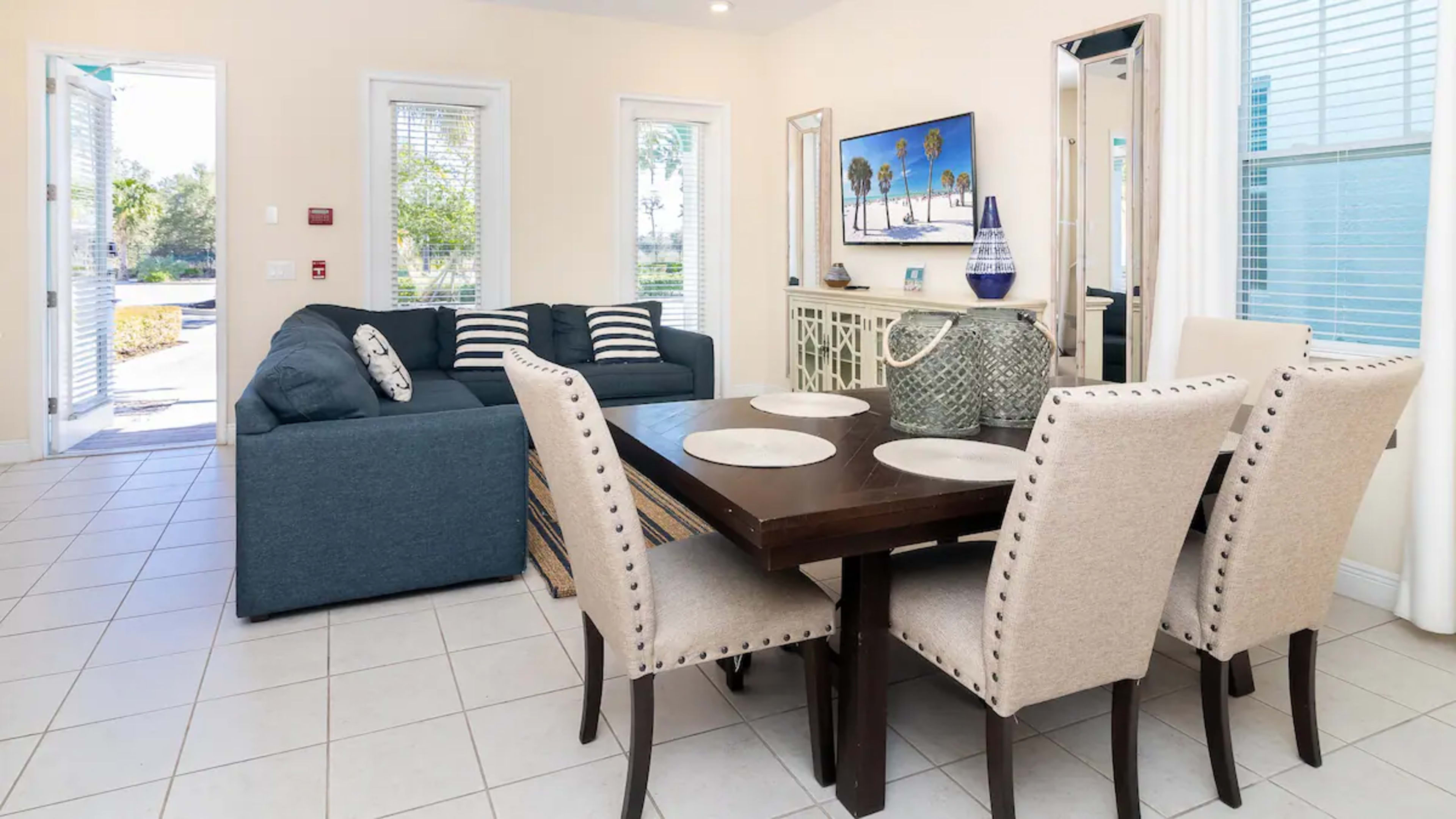 A living and dining area featuring a blue sofa, a dining table with beige chairs, and large windows providing natural light.