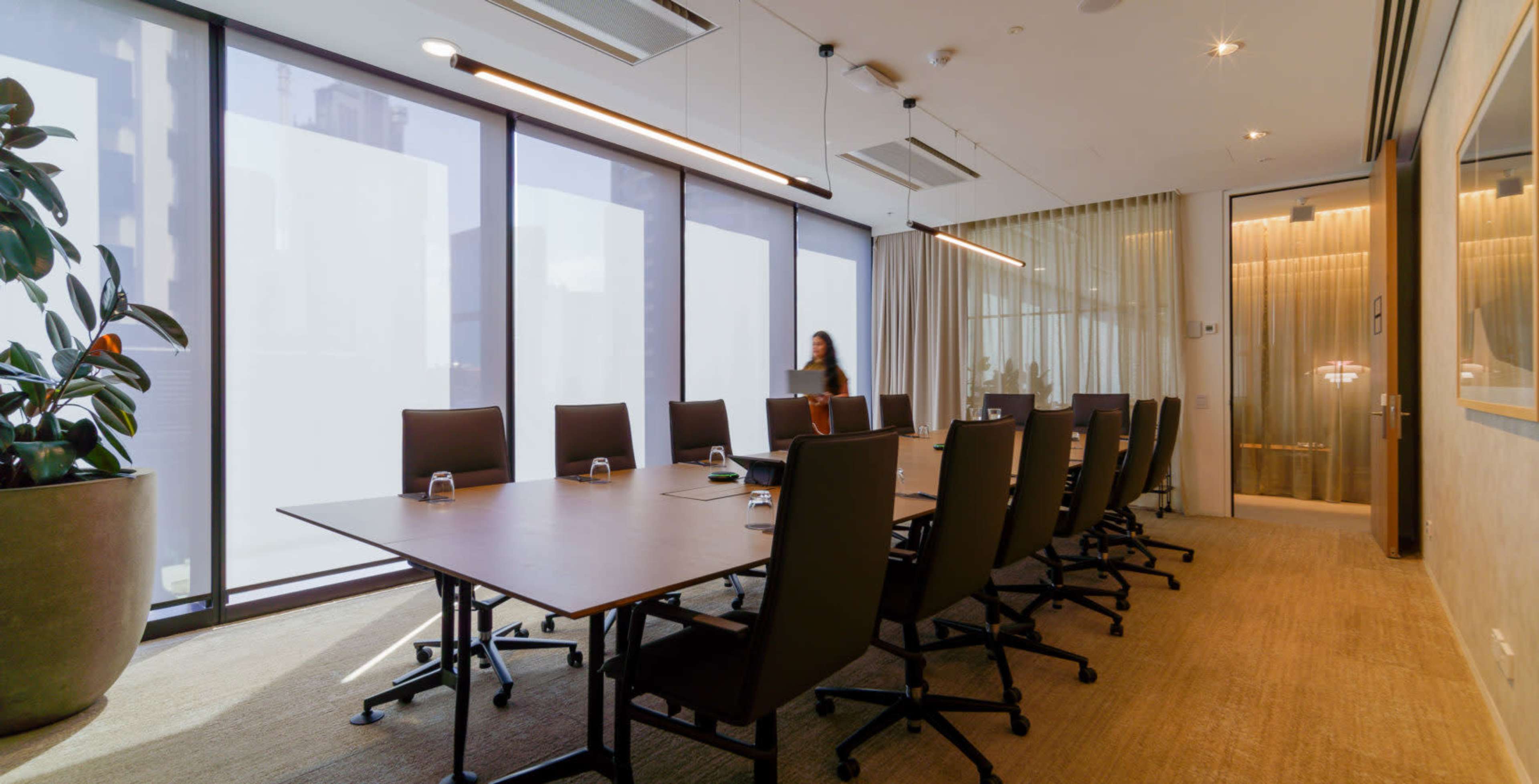 A modern conference room features a long wooden table surrounded by black chairs and large windows with sheer curtains.