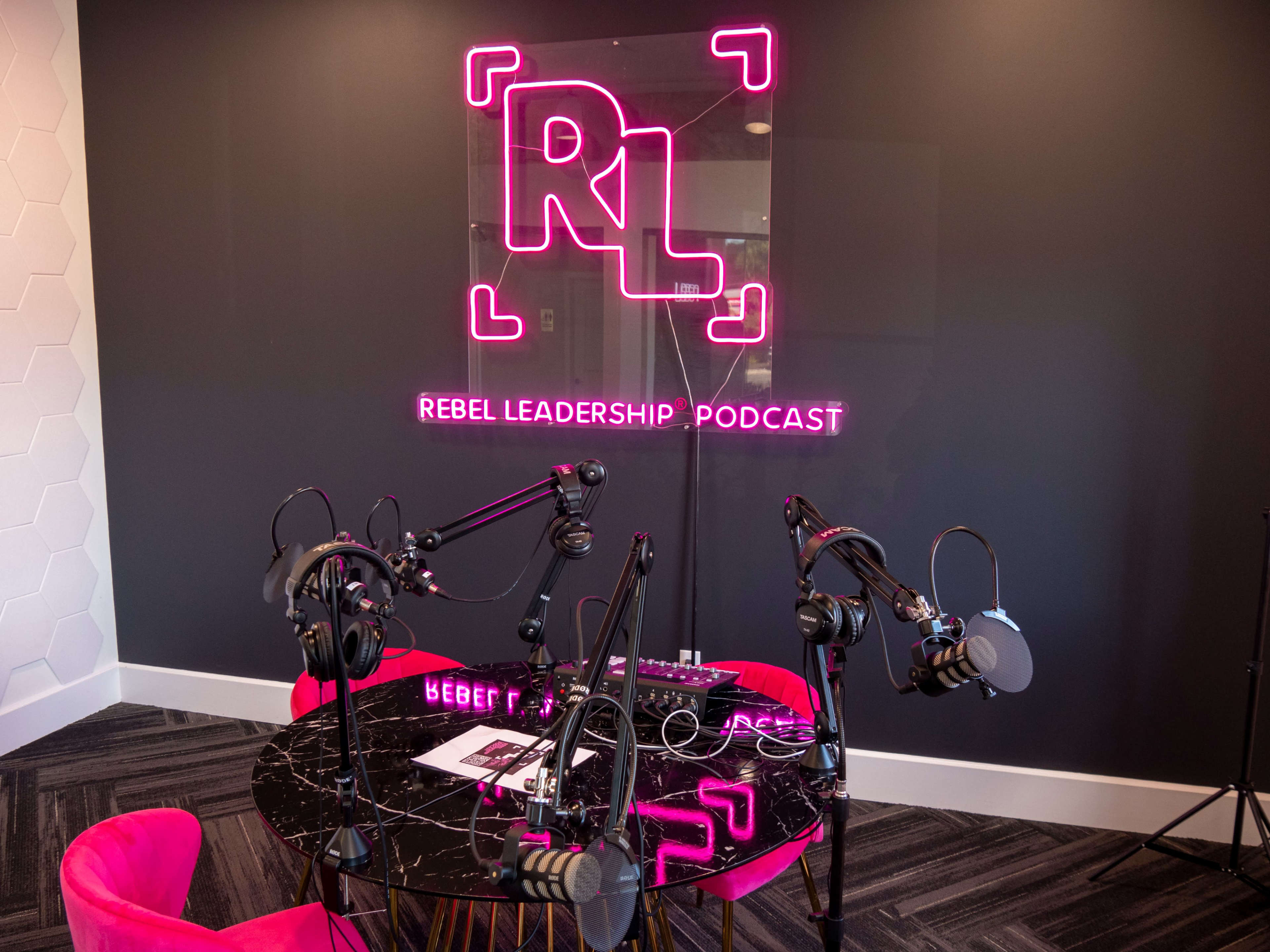 A podcasting studio featuring a black table with multiple microphones and headphones, illuminated by a neon sign that reads "REBEL LEADERSHIP PODCAST."