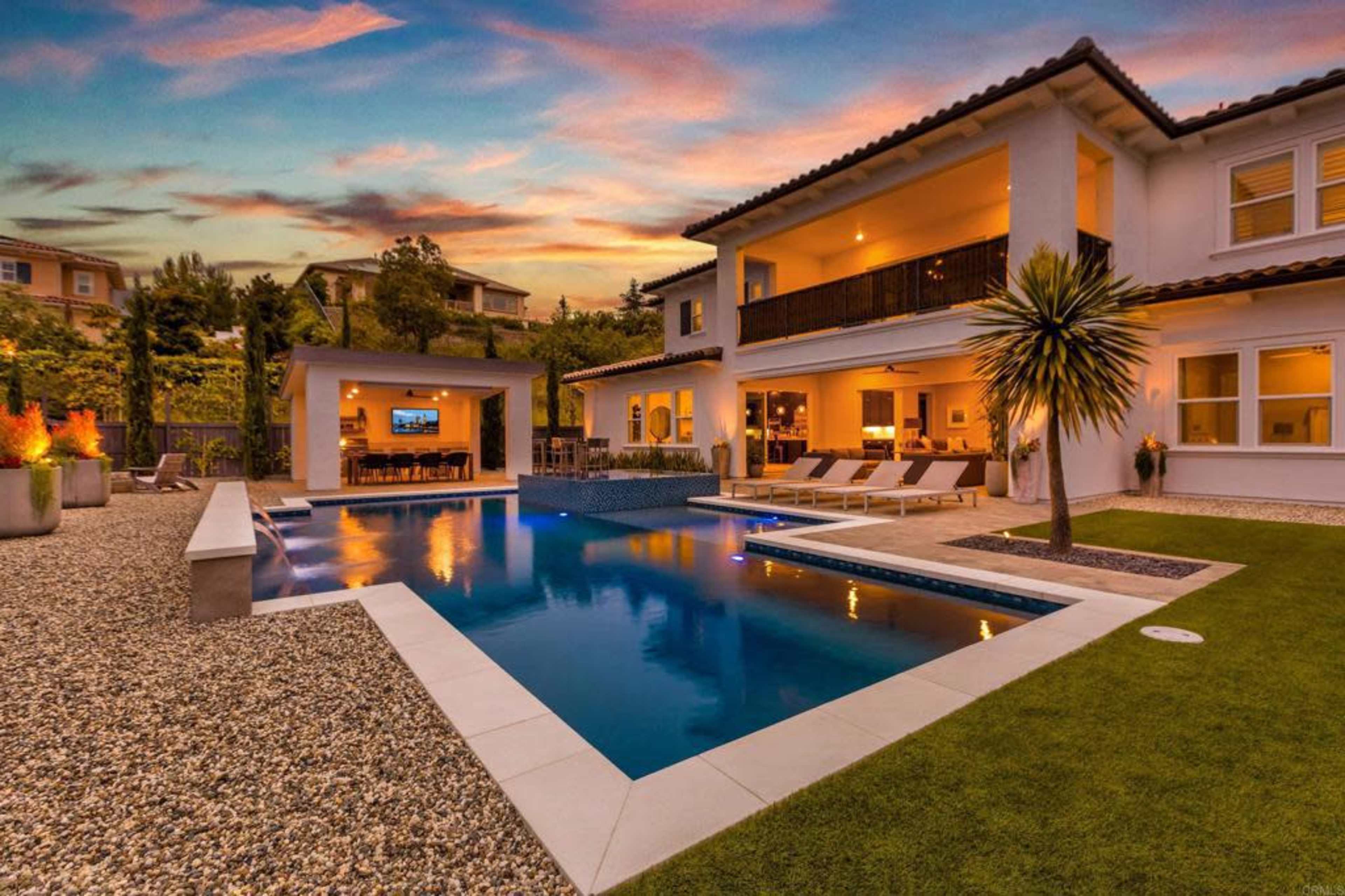 A modern backyard with a swimming pool, lounge chairs, and a spacious home illuminated at sunset.