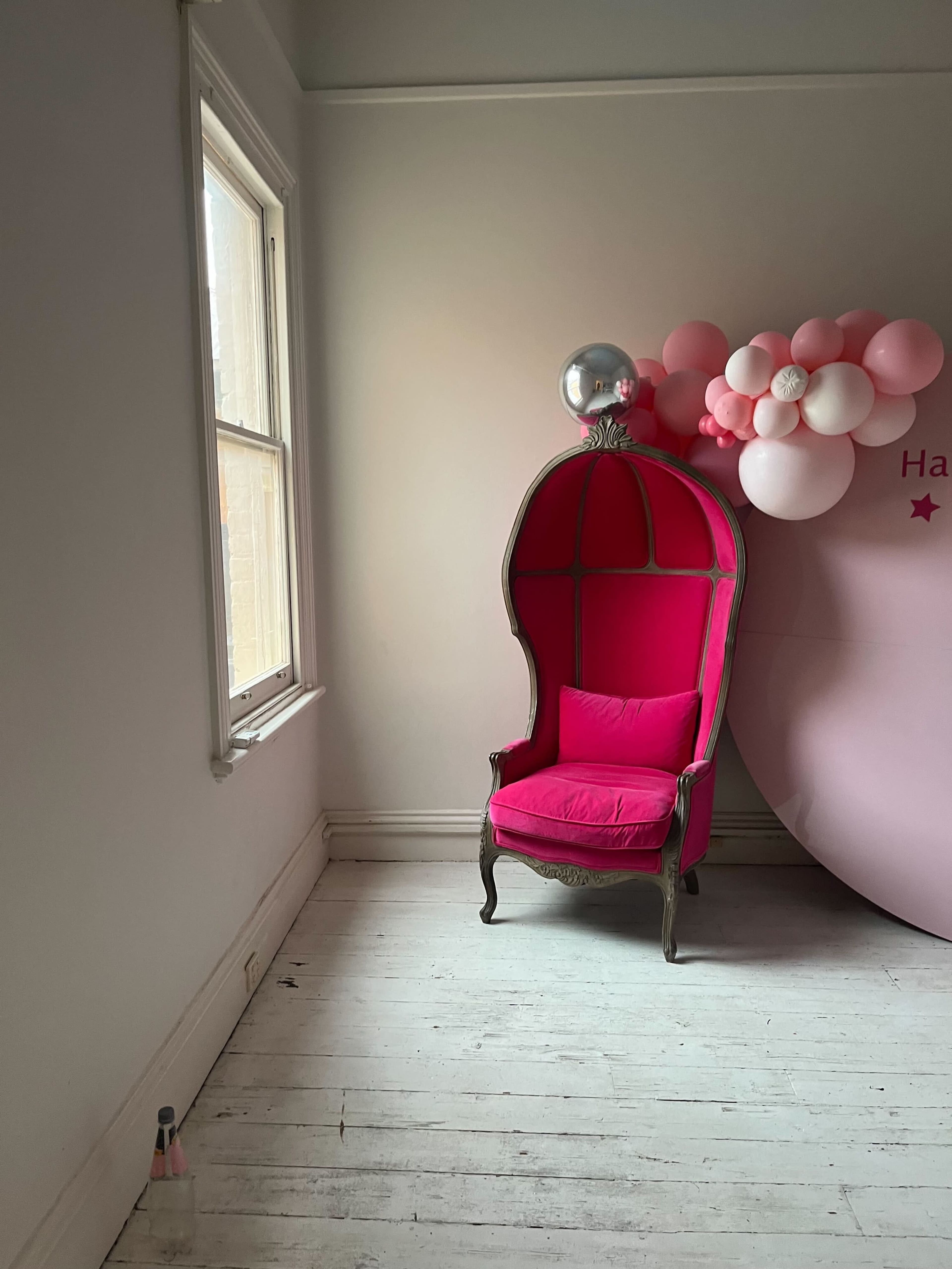 A pink ornate chair positioned beside a window, complemented by a cluster of pink and white balloons against a wall.
