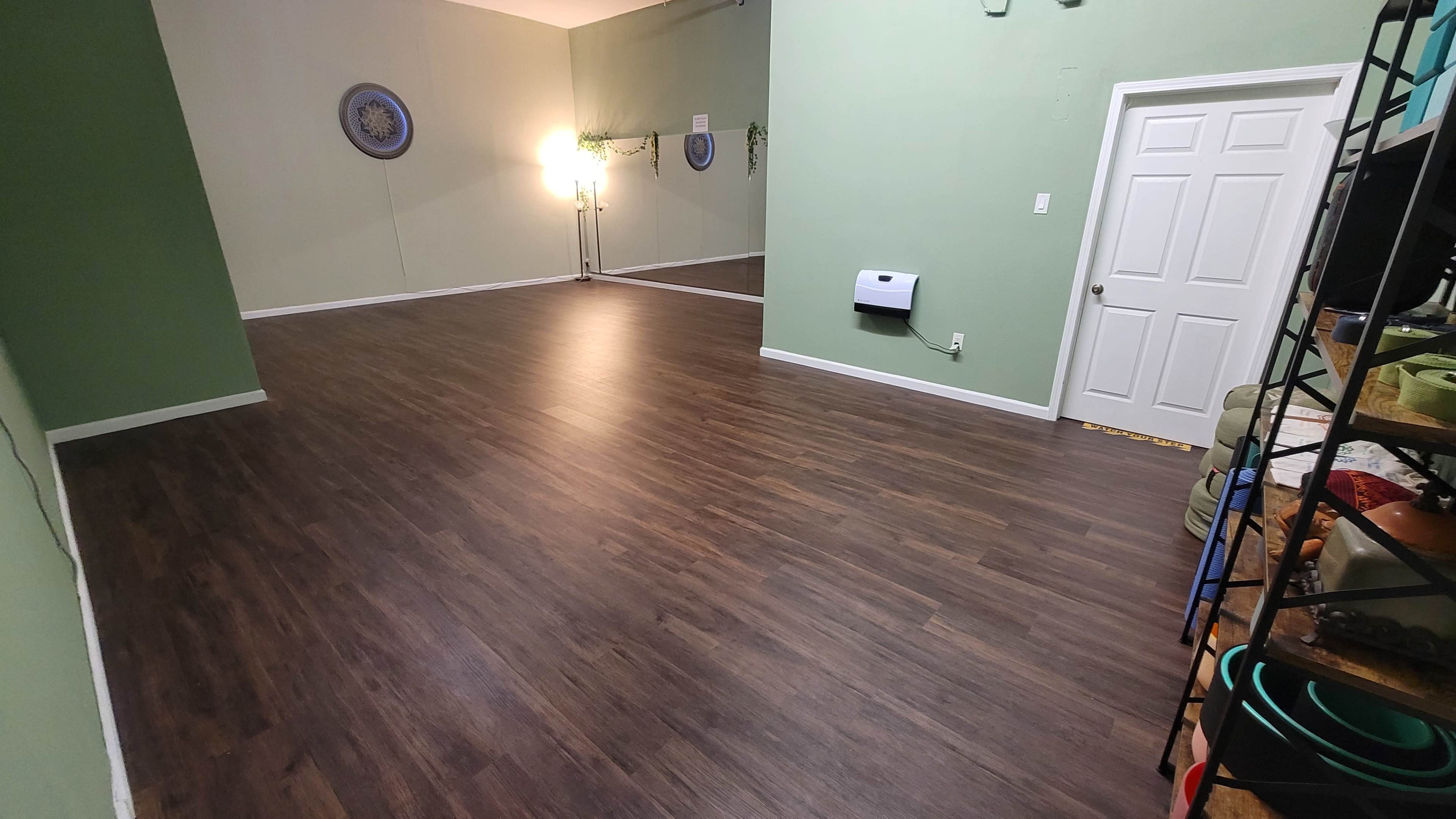 The image shows a spacious room with wooden floors, green walls, a mirror, and a closed door on one side.