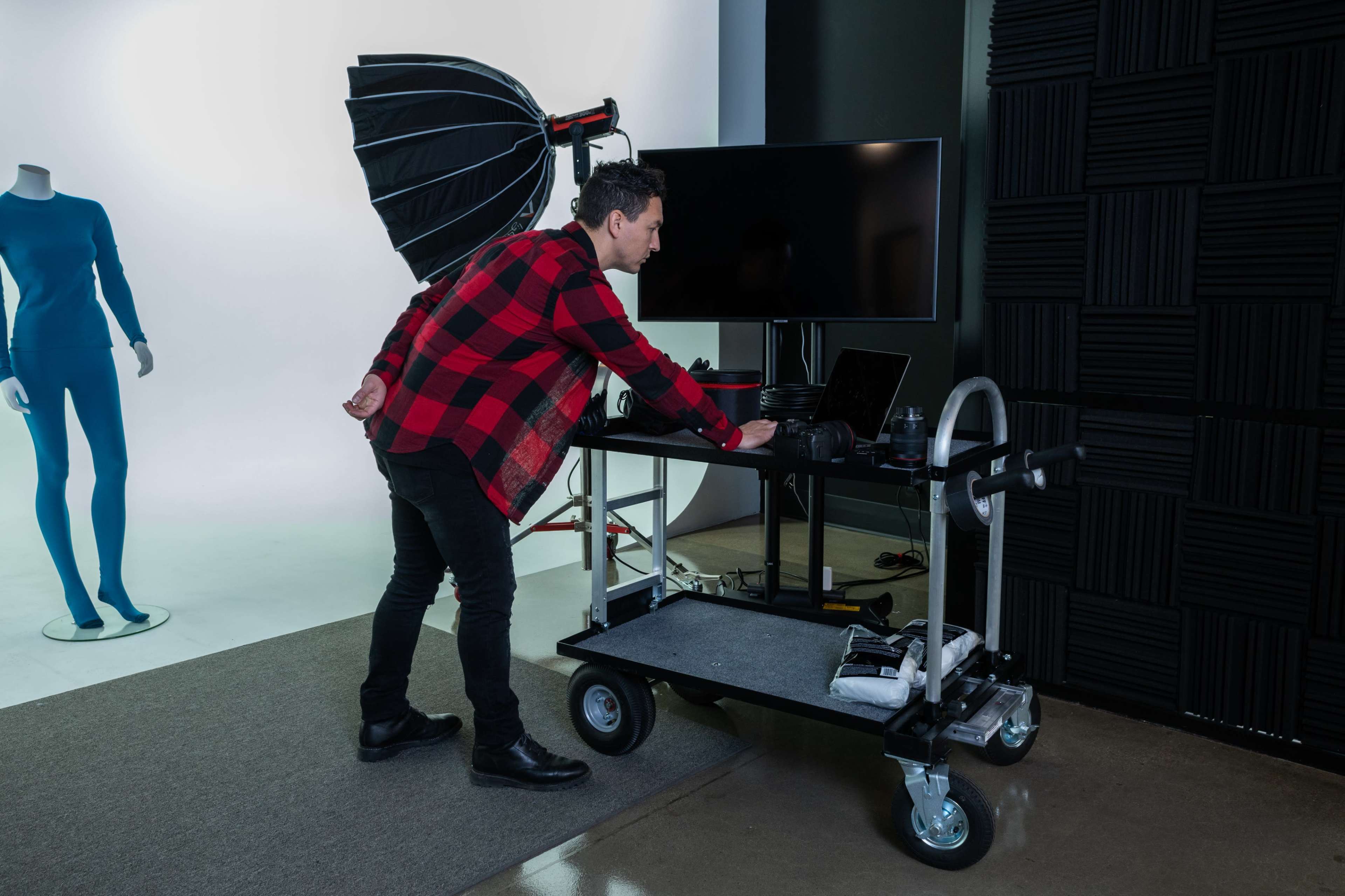 A person in a plaid shirt adjusts a laptop on a cart in a studio setting with a mannequin and lighting equipment in the background.