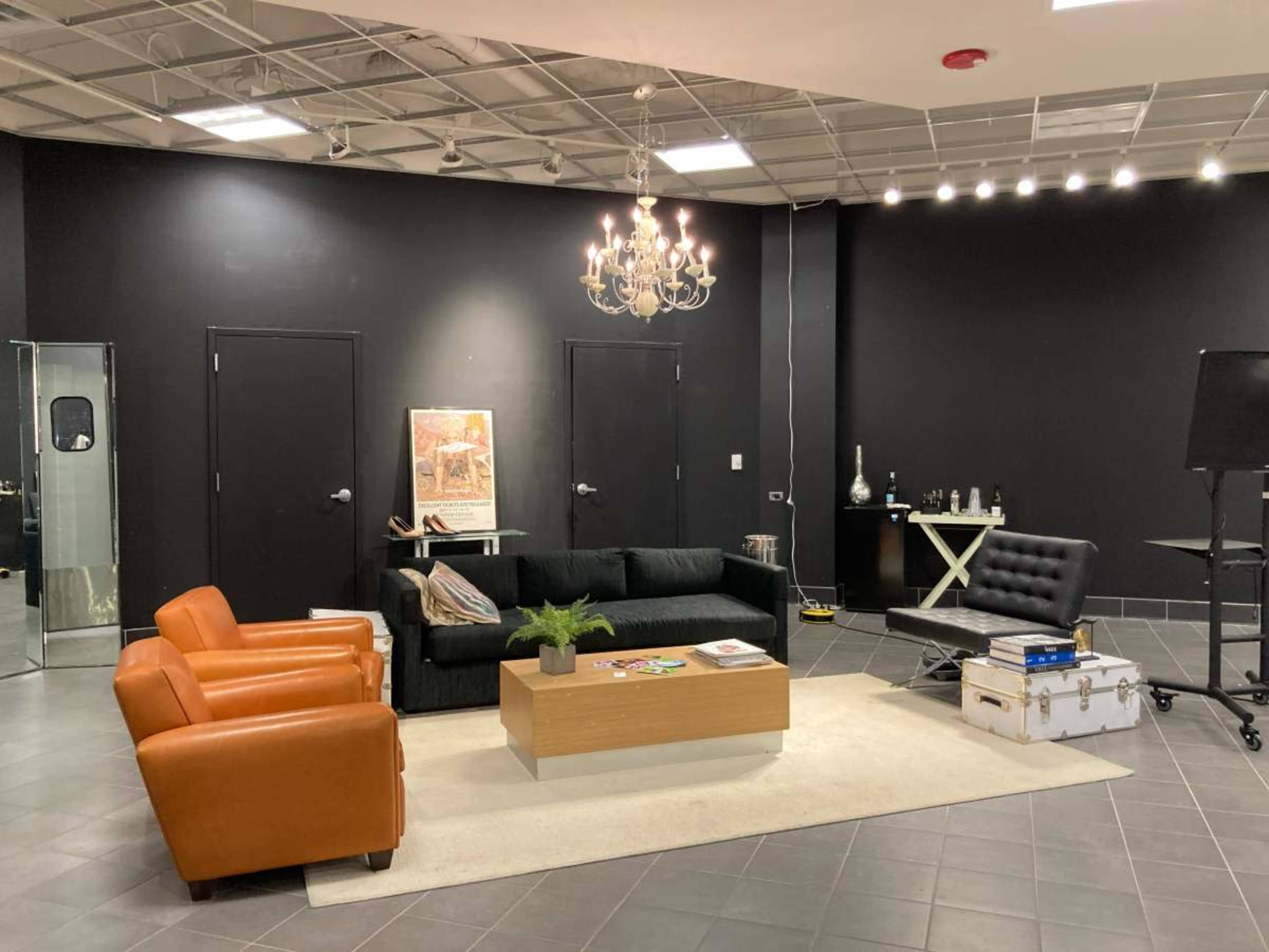 The image shows a modern lounge area with a black wall, two orange chairs, a black couch, a coffee table, and a chandelier overhead.