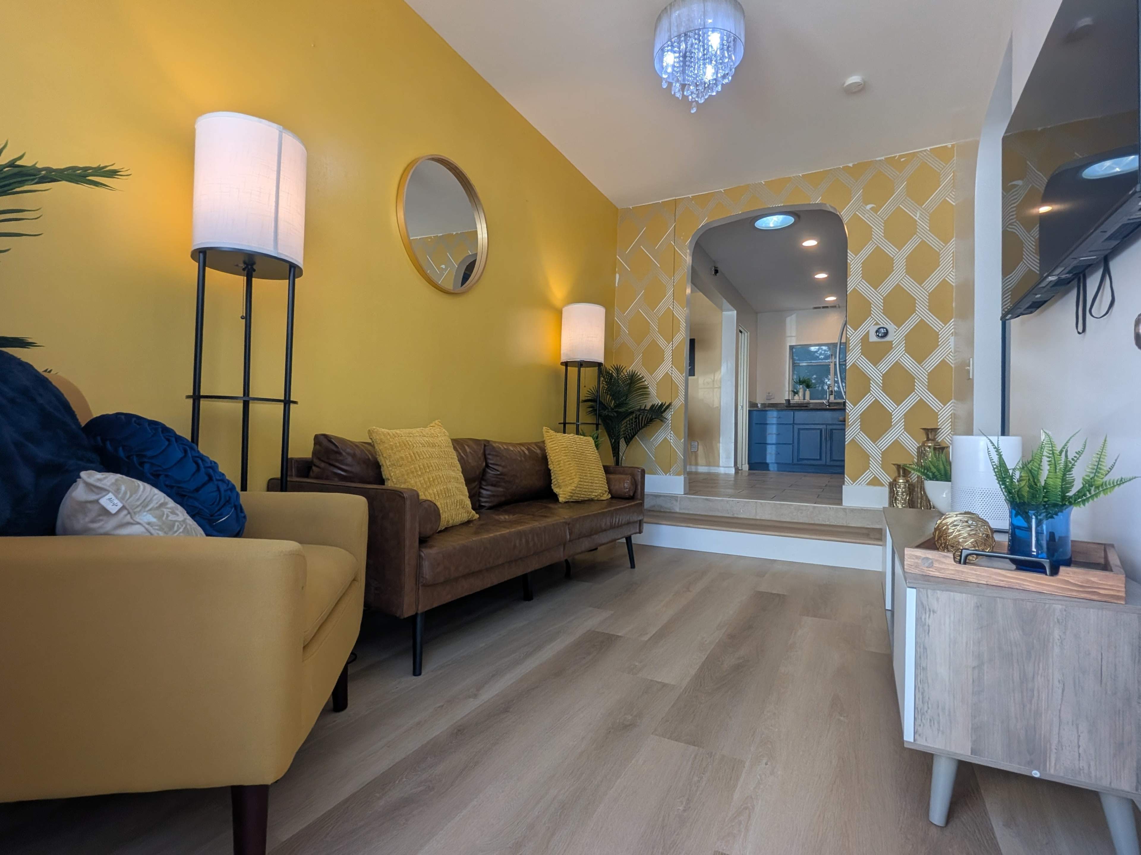 The image shows a brightly colored living room with yellow walls, a brown leather sofa, decorative pillows, and an archway leading to a kitchen area.