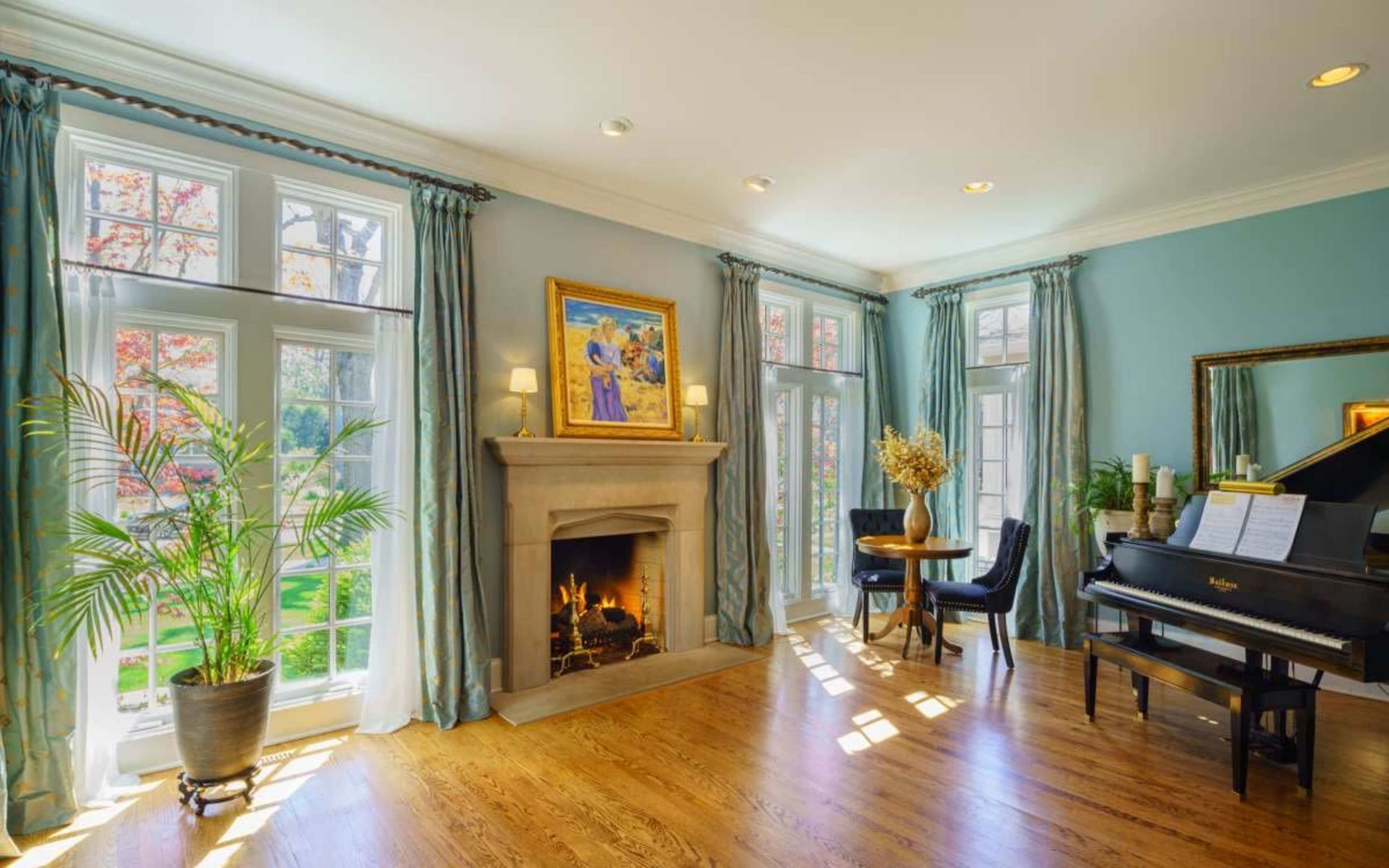 The image shows a well-lit living room featuring a fireplace, a grand piano, a small dining table, and large windows with blue curtains.