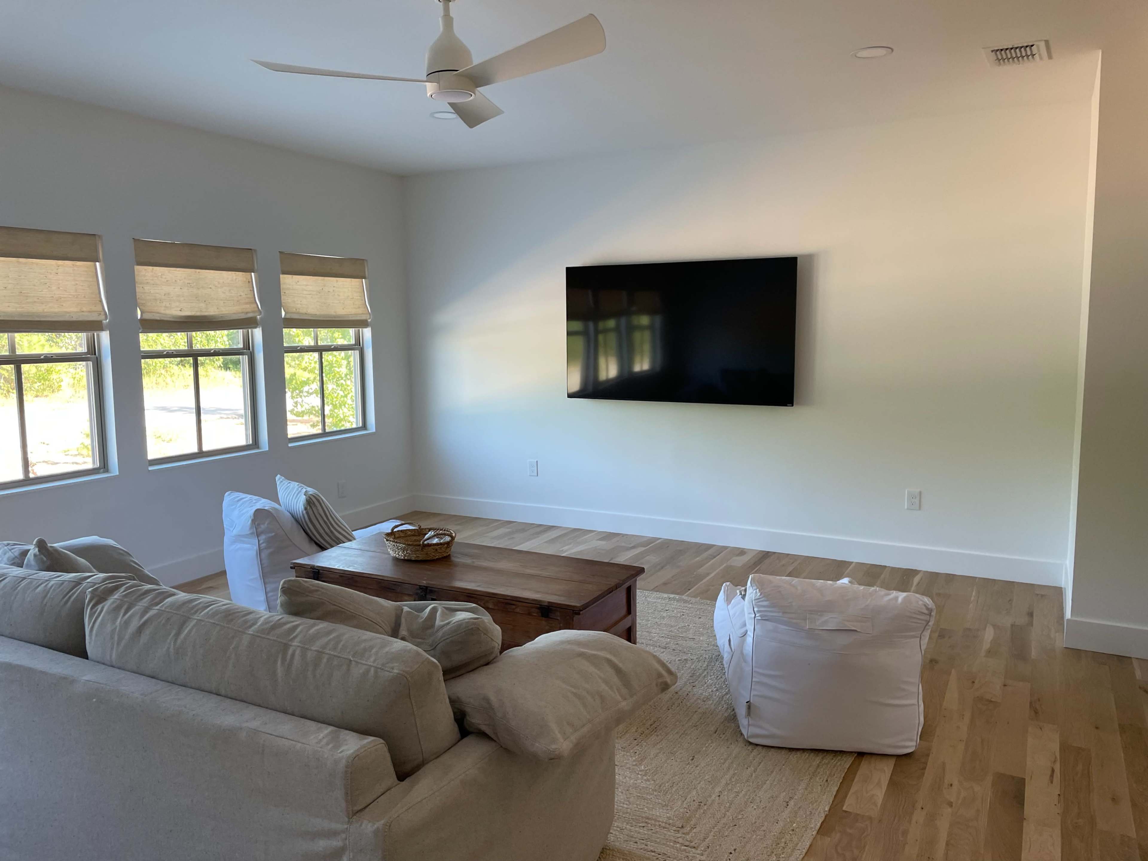 A light-filled living room features a sofa, a wooden coffee table, and a large flat-screen TV mounted on the wall.
