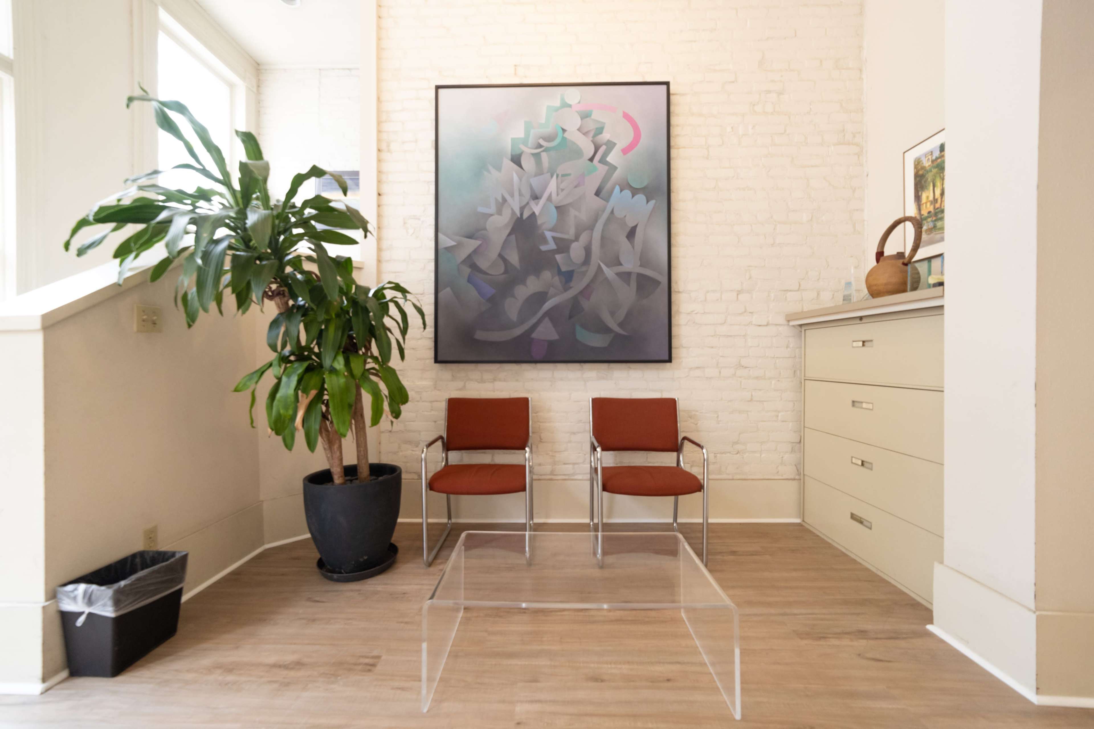 A modern interior space with two metal chairs, a clear acrylic coffee table, a large abstract painting on the wall, and a potted plant in a corner.