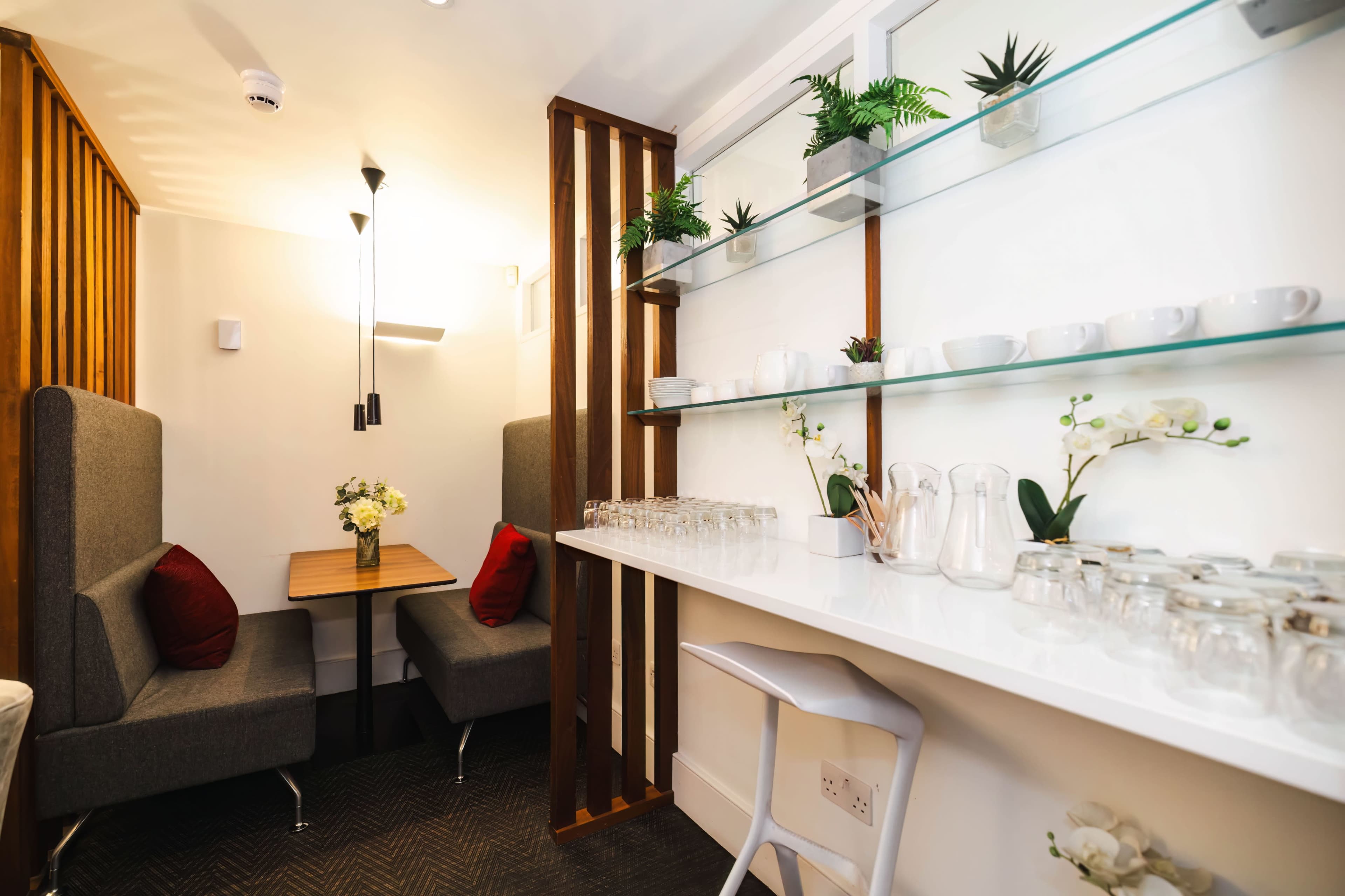A small seating area with two gray booths, a wooden table, and a white counter displaying glassware and plants against a light-colored wall.